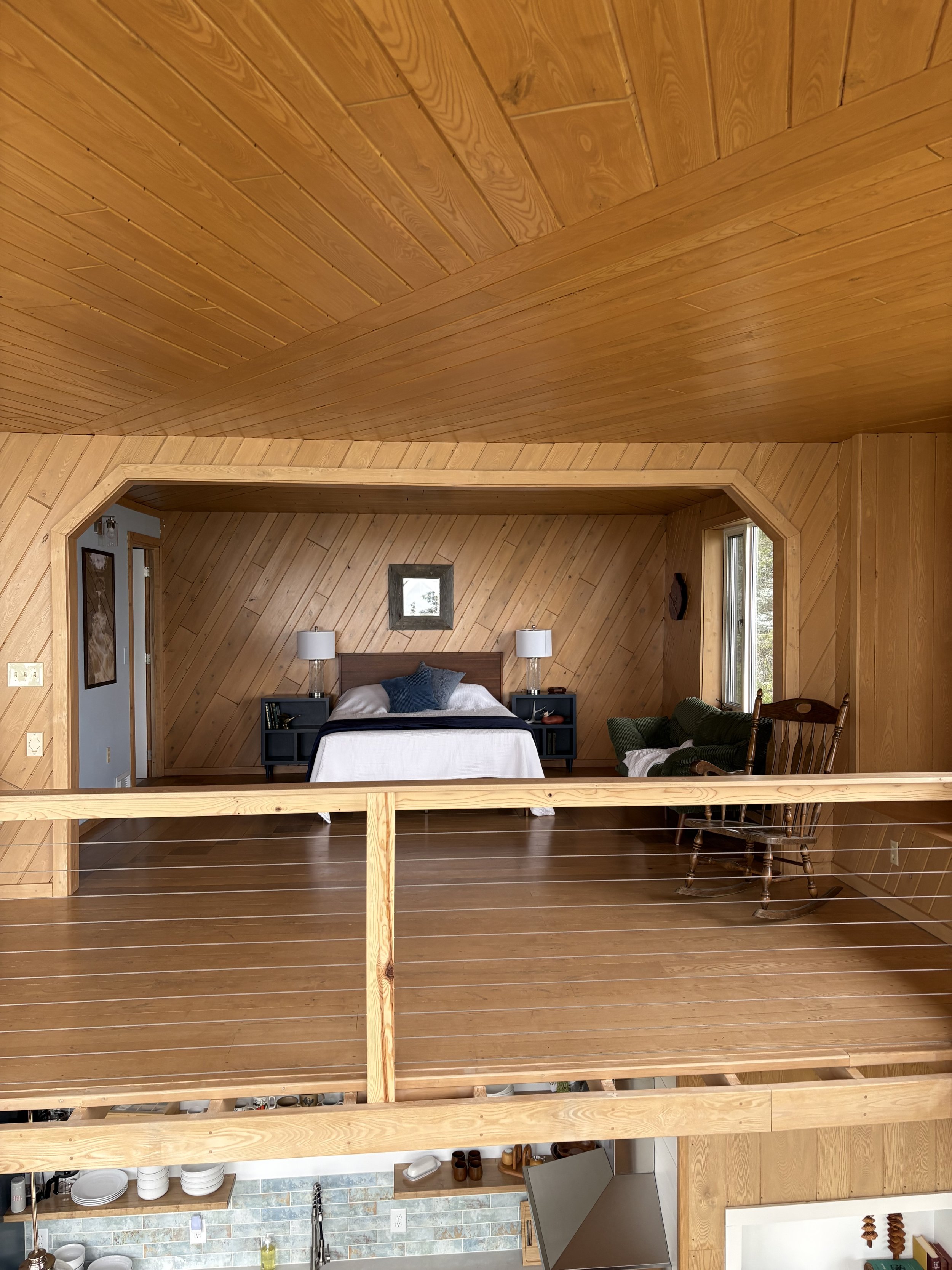 Interior of a house with wood-paneled walls and ceiling, containing a cozy bedroom with a bed flanked by nightstands and lamps, and a rocking chair near a window, with a view into the kitchen below.