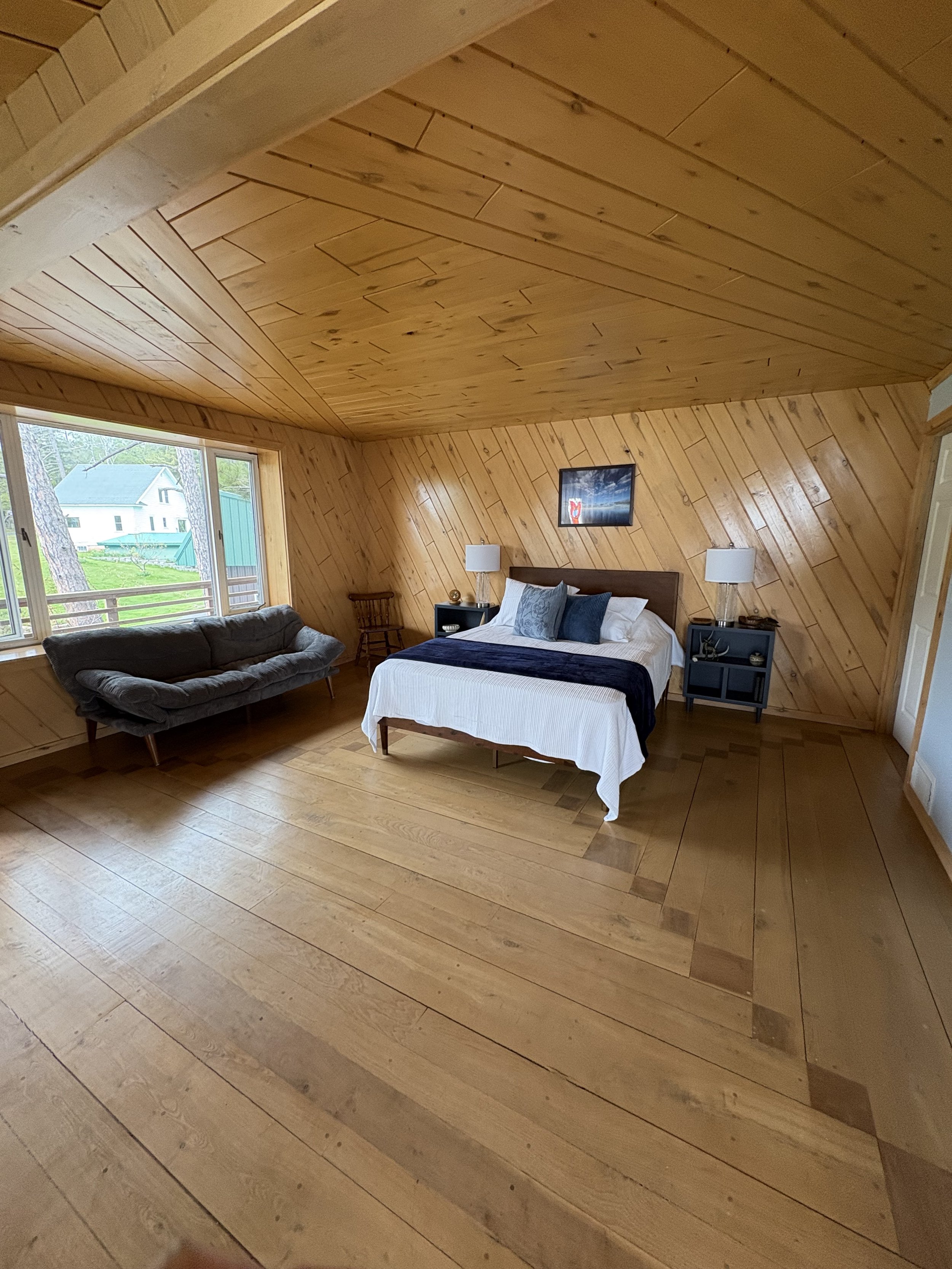 A room with wooden interior walls and ceiling, a window showing trees and a house outside, a gray couch, a bed with white bedding, two lamps on black side tables, and a framed picture on the wall.