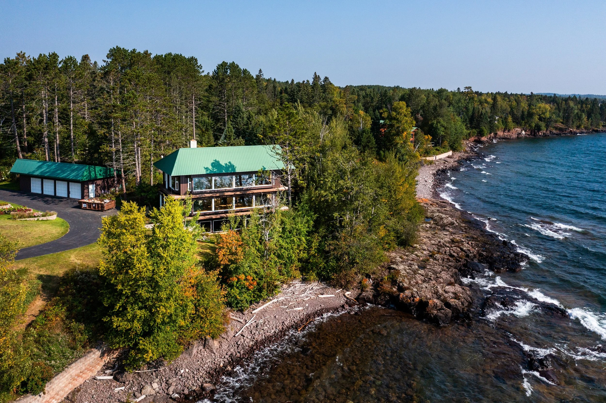 Aerial view of a house with a green roof along a rocky shoreline, surrounded by dense green forest and clear blue water.