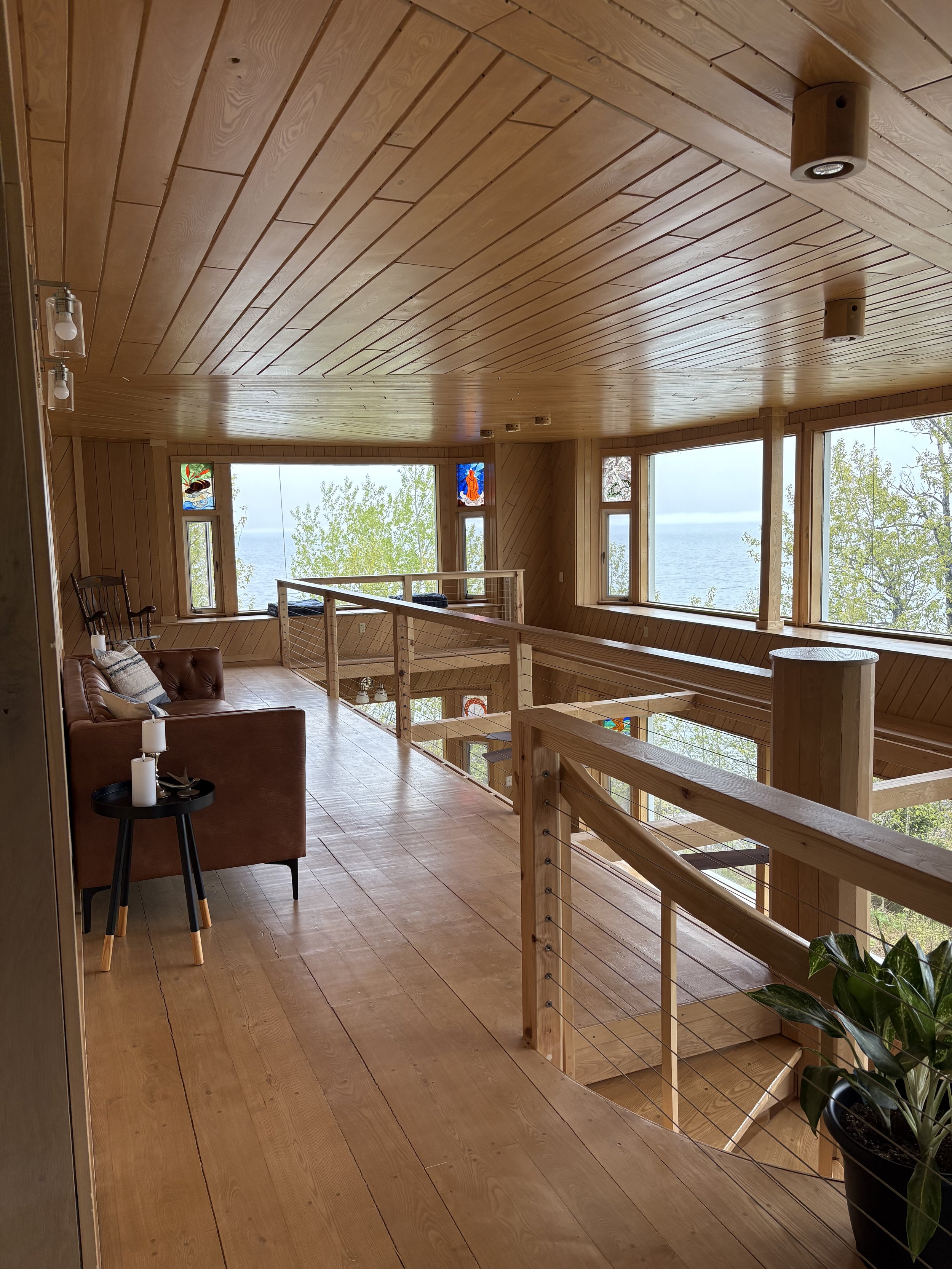 Indoor view of a wooden room with large windows overlooking water. There is a brown leather sofa, a small black table with candles, and potted plant. Wooden ceiling with installed lights.