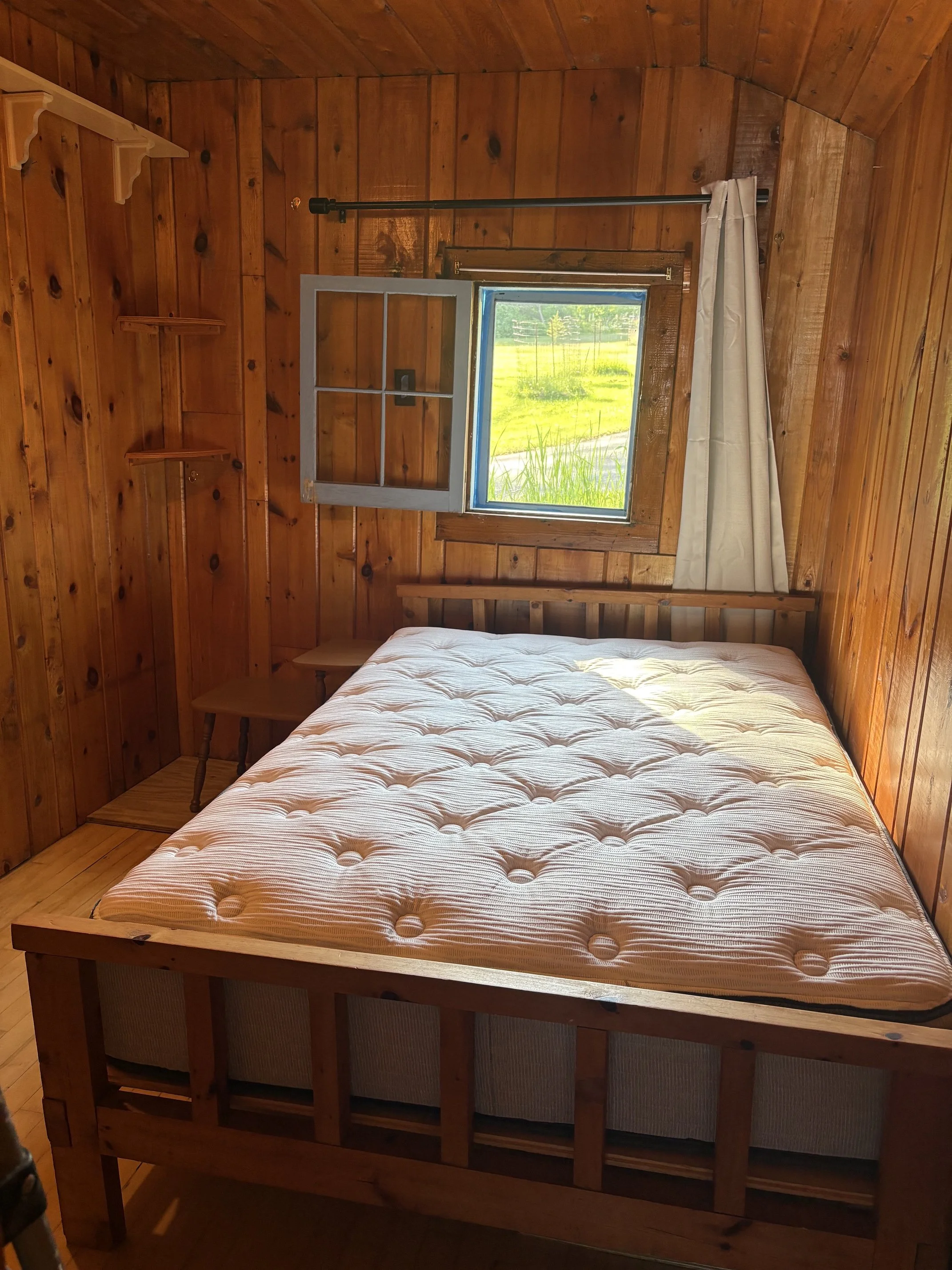 A bedroom with wooden walls and ceiling, featuring a window with a white curtain partially pulled to the side, showing green grass and trees outside. There is a bed with a beige mattress, and a small wooden side table next to it.
