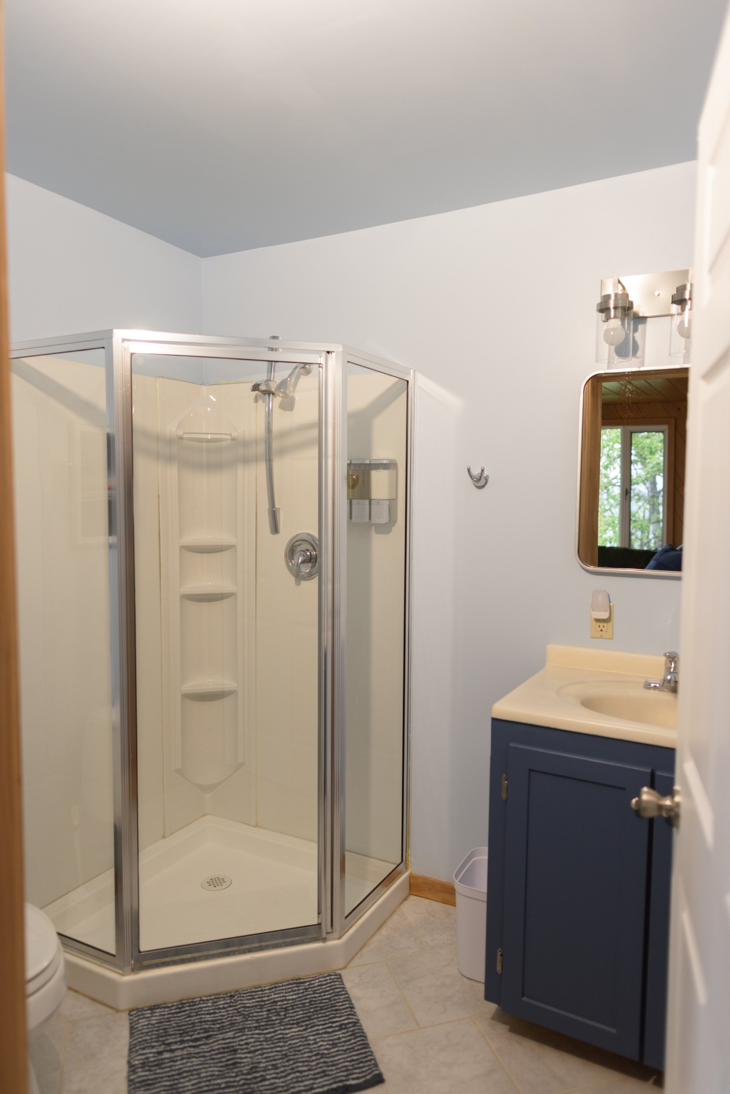 Small bathroom featuring a corner shower stall with a glass door, a vanity with a blue cabinet, a mirror, and a window with greenery outside.