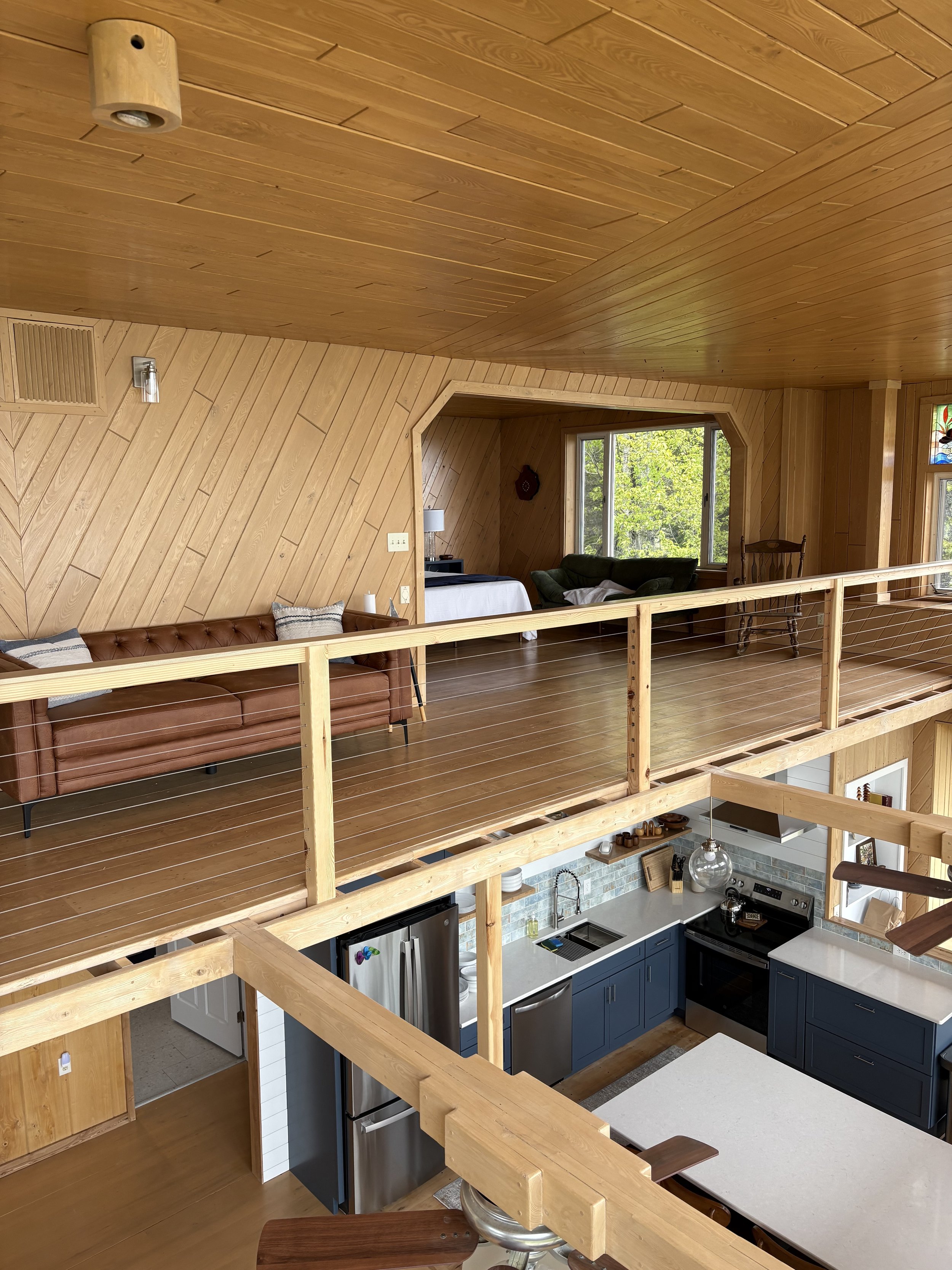Interior view of a multi-level house with wooden walls and ceilings, featuring a living area with a leather couch, a kitchen with blue cabinets, and a large window showing lush green trees outside.