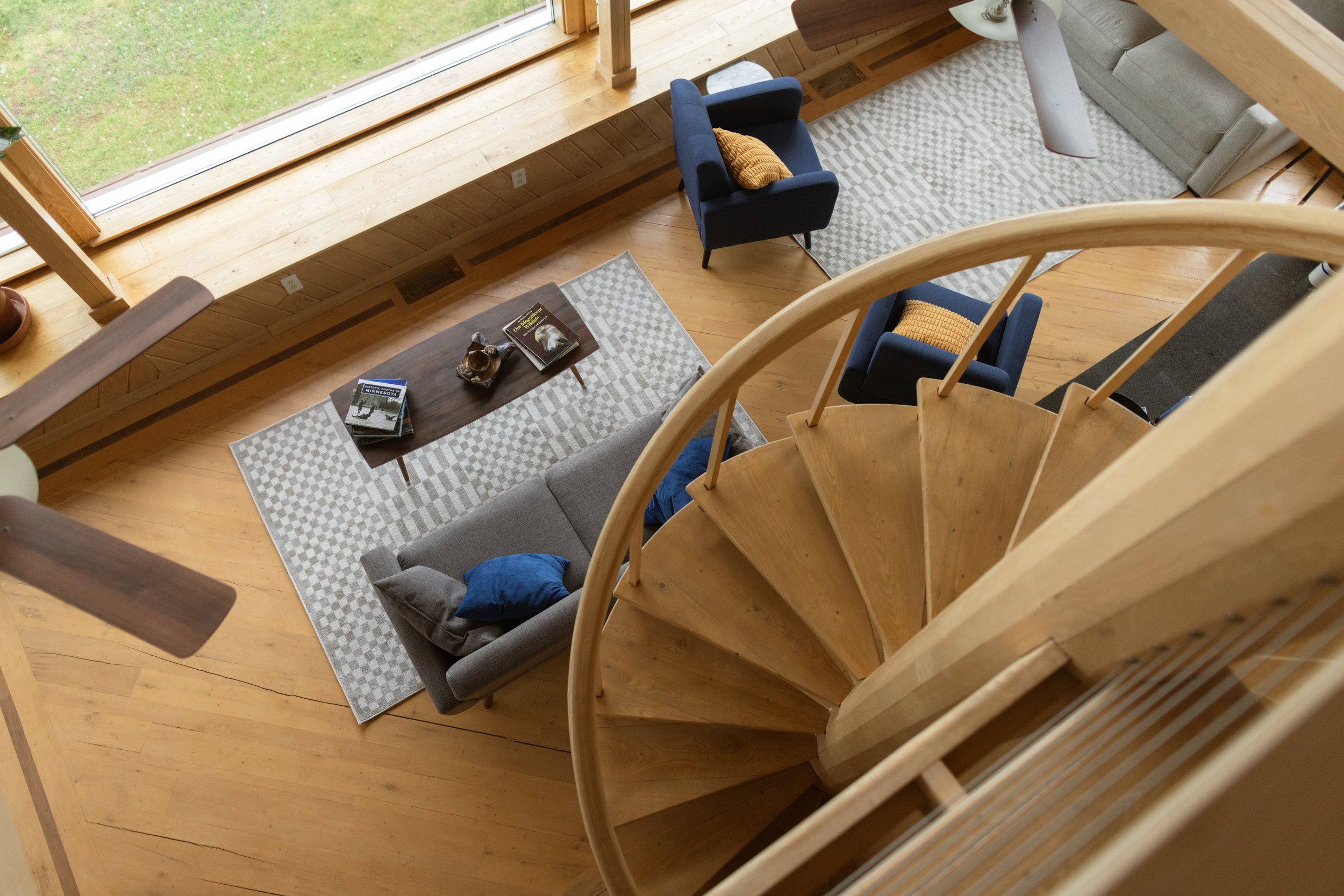 Overhead view of a living room with wooden floors, a large window overlooking a green yard, a gray sofa with pillows, two blue armchairs with yellow pillows, a wooden coffee table with books and decorative objects, and a light-colored area rug.