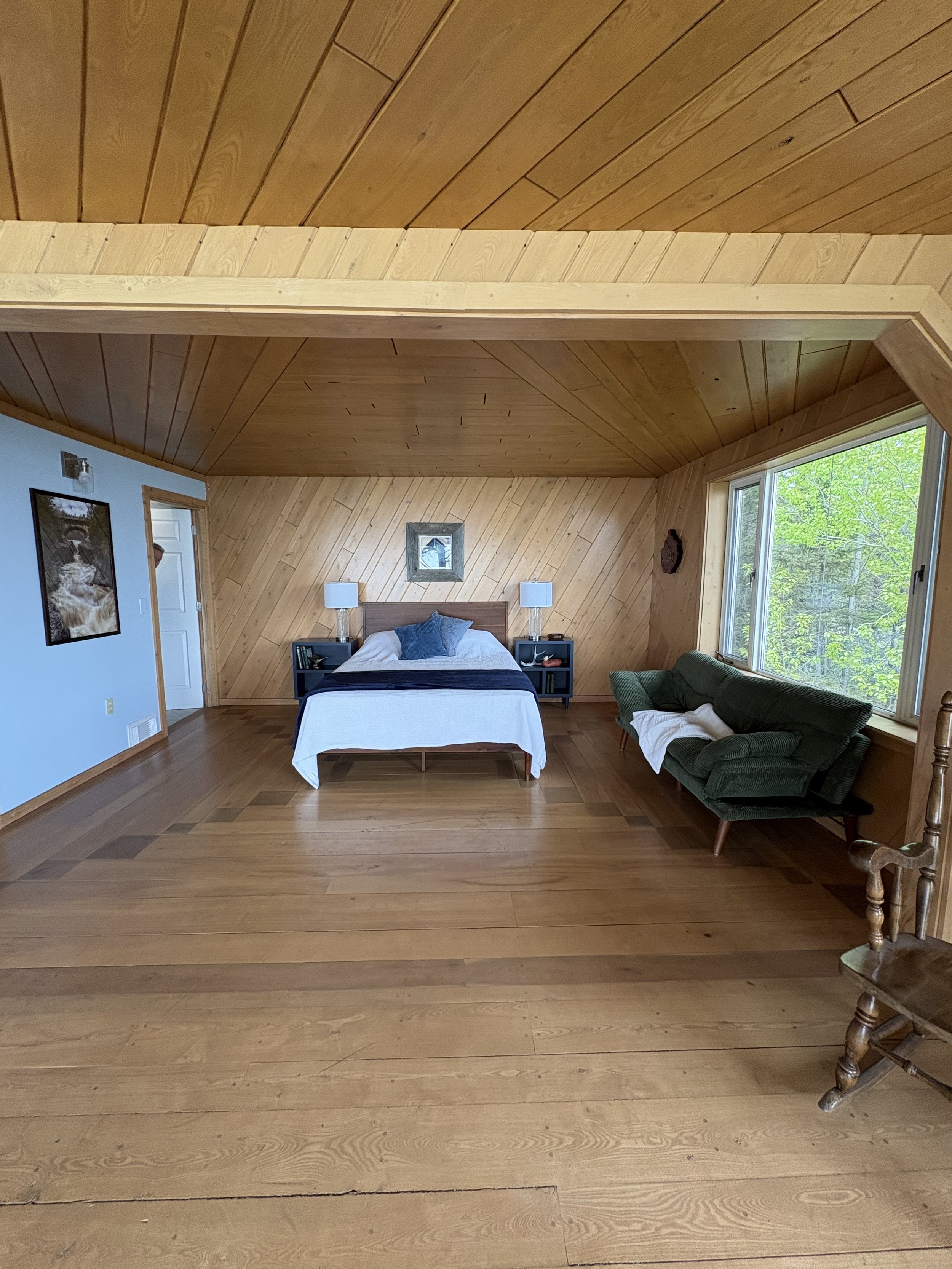 A bedroom with wooden paneled walls and ceiling, a bed with pillows and a white cover, nightstands with lamps, a window showing green trees, a green armchair, a framed picture on the wall, and a wooden rocking chair.