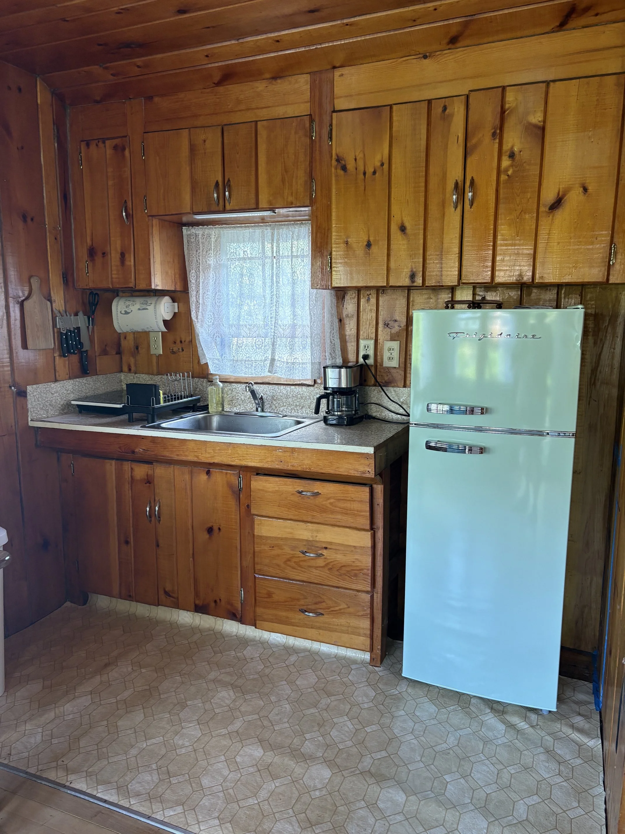 Kitchen with wooden cabinets, a window with lace curtains, a white vintage refrigerator, a coffee maker, and a sink with dish soap and a drying rack.