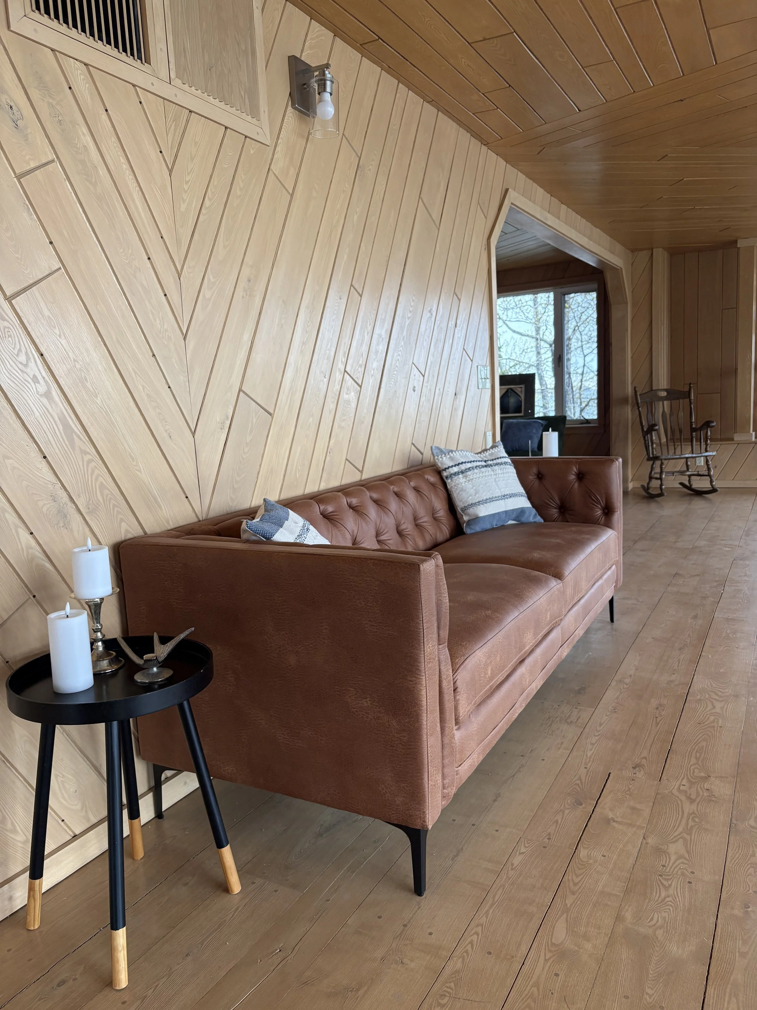 Living room with wood-paneled walls and ceiling, a brown leather sofa with striped pillows, a small black side table with candles, and a view of a forest through a large window.