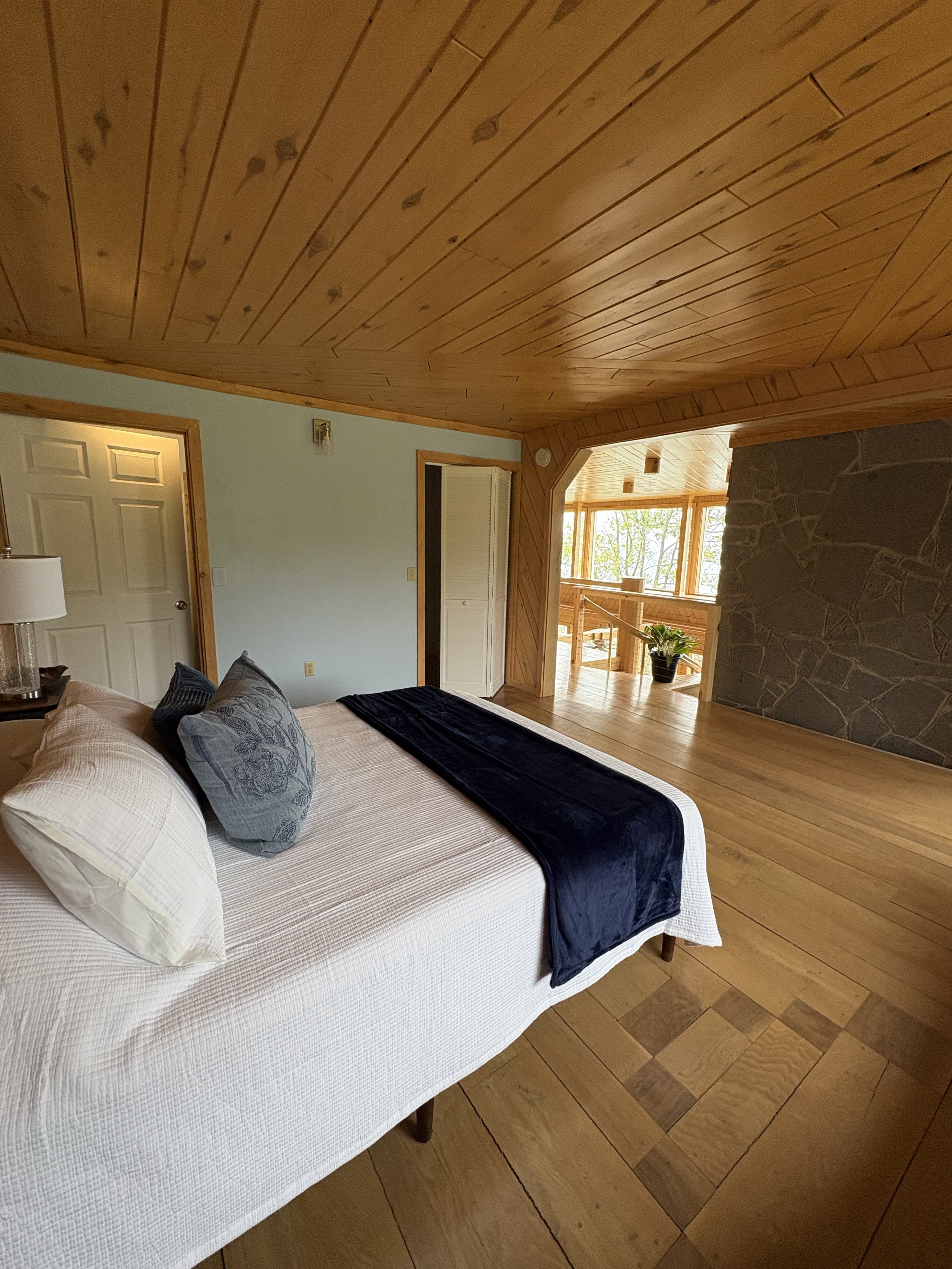 Interior of a room with a bed, wooden ceiling and floor, a stone fireplace, and windows with trees outside.