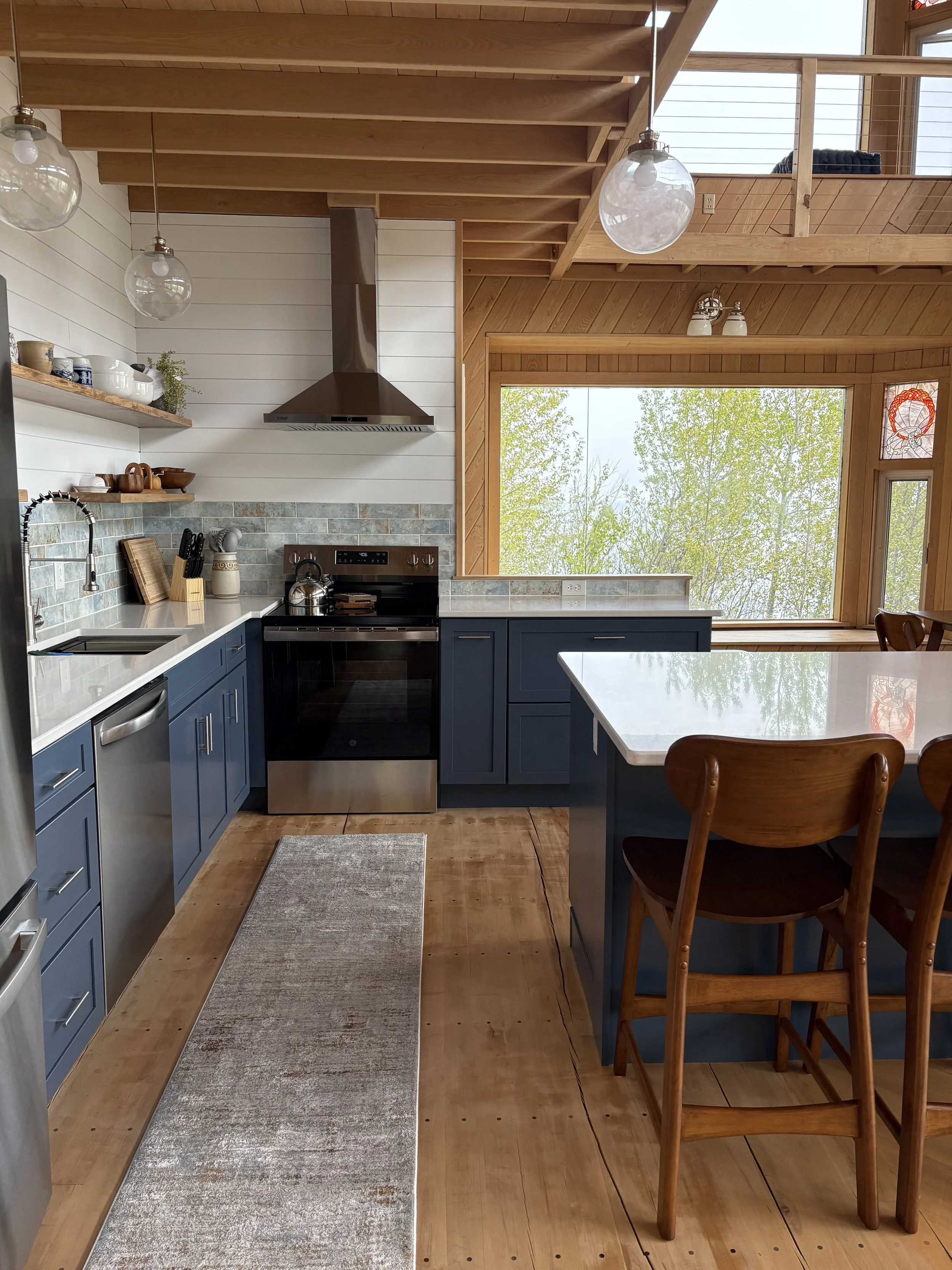 A modern kitchen with blue cabinets, a white countertop, a stainless steel oven, and a window overlooking trees. There are two wooden chairs at a white kitchen island and open wooden shelves with dishes and decorative items. The ceiling has wooden pa