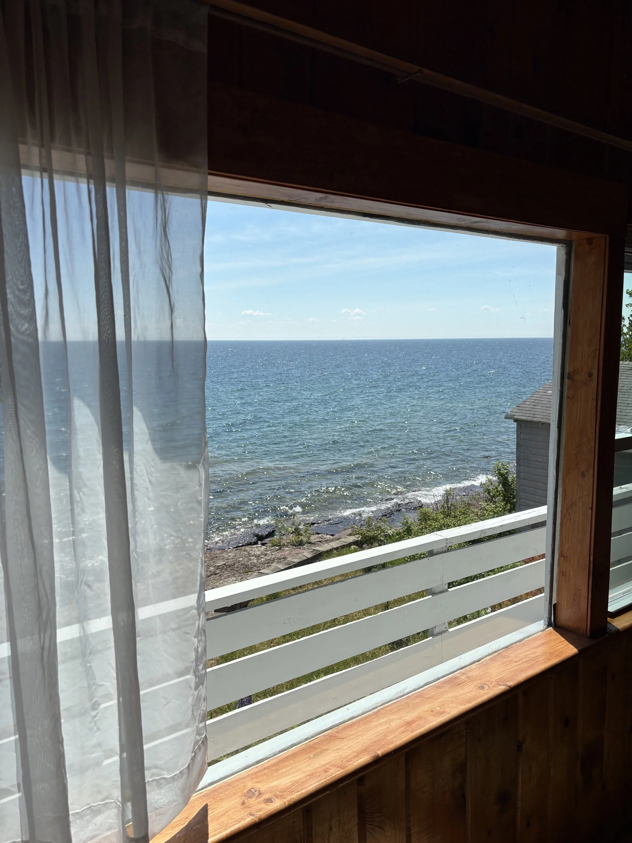 Lake view through a wooden-framed window. The window has sheer white curtains and a white railing outside, with rocks and water below.