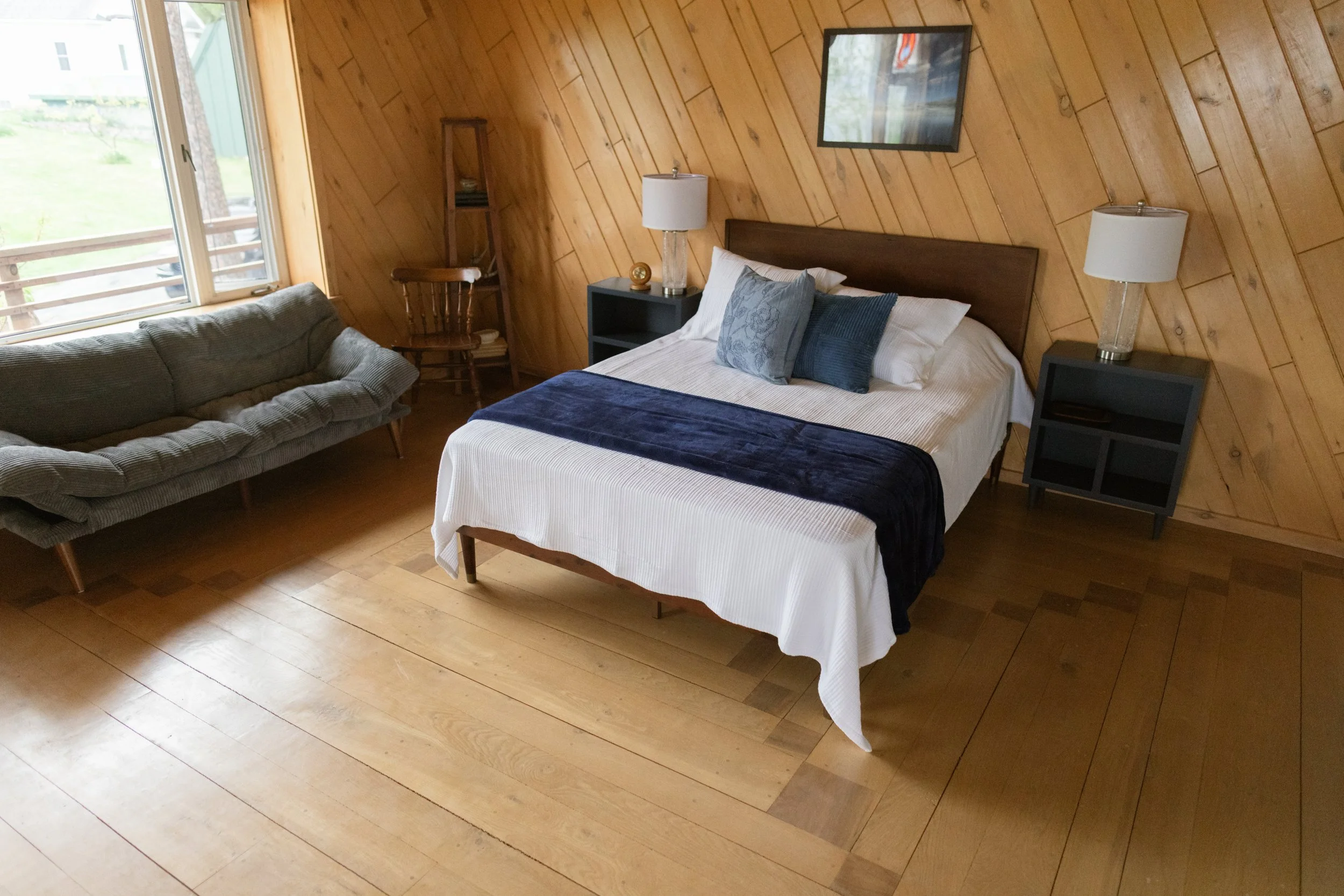 A bedroom with wooden panel walls, a bed with white linens and blue pillows, dark nightstands with lamps, a wooden chair, a window with a view of a grassy yard, and a wall mirror.