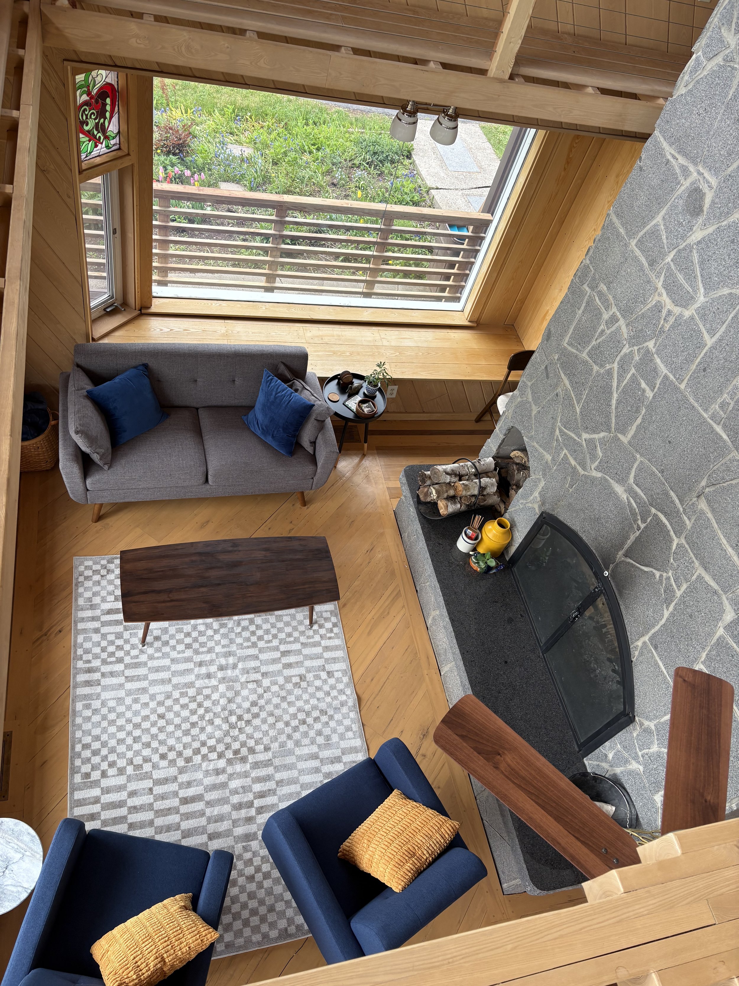 Living room with wooden floors and walls, a gray sofa with blue pillows, a black coffee table, a beige and white checkered rug, a stone fireplace with logs, a large window showing a garden, and a sitting area with a wooden chair.