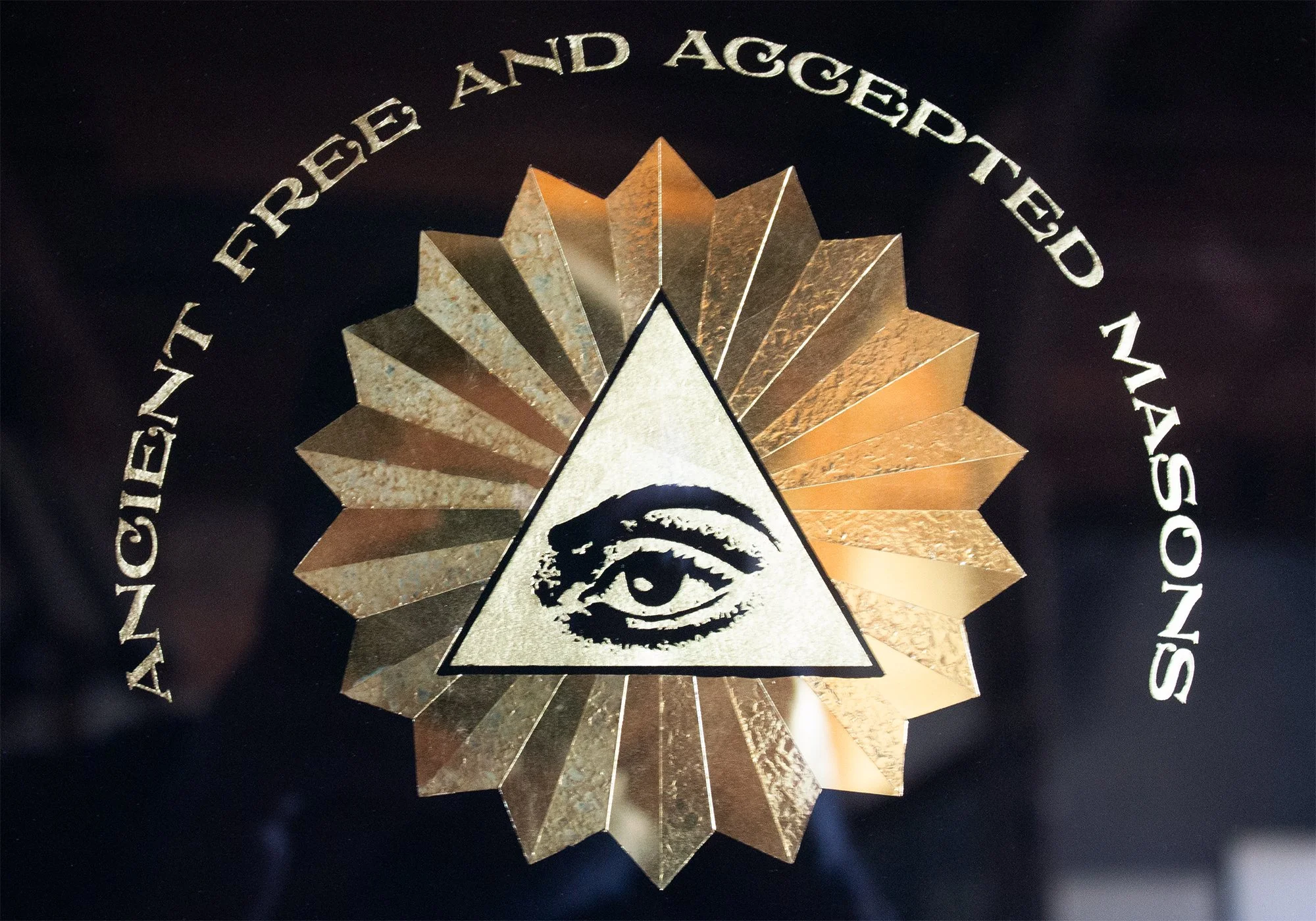 A logo with a gold starburst design, a black triangular eye symbol in the center, and the words 'Ancient Free and Accepted Masons' curved above the starburst.