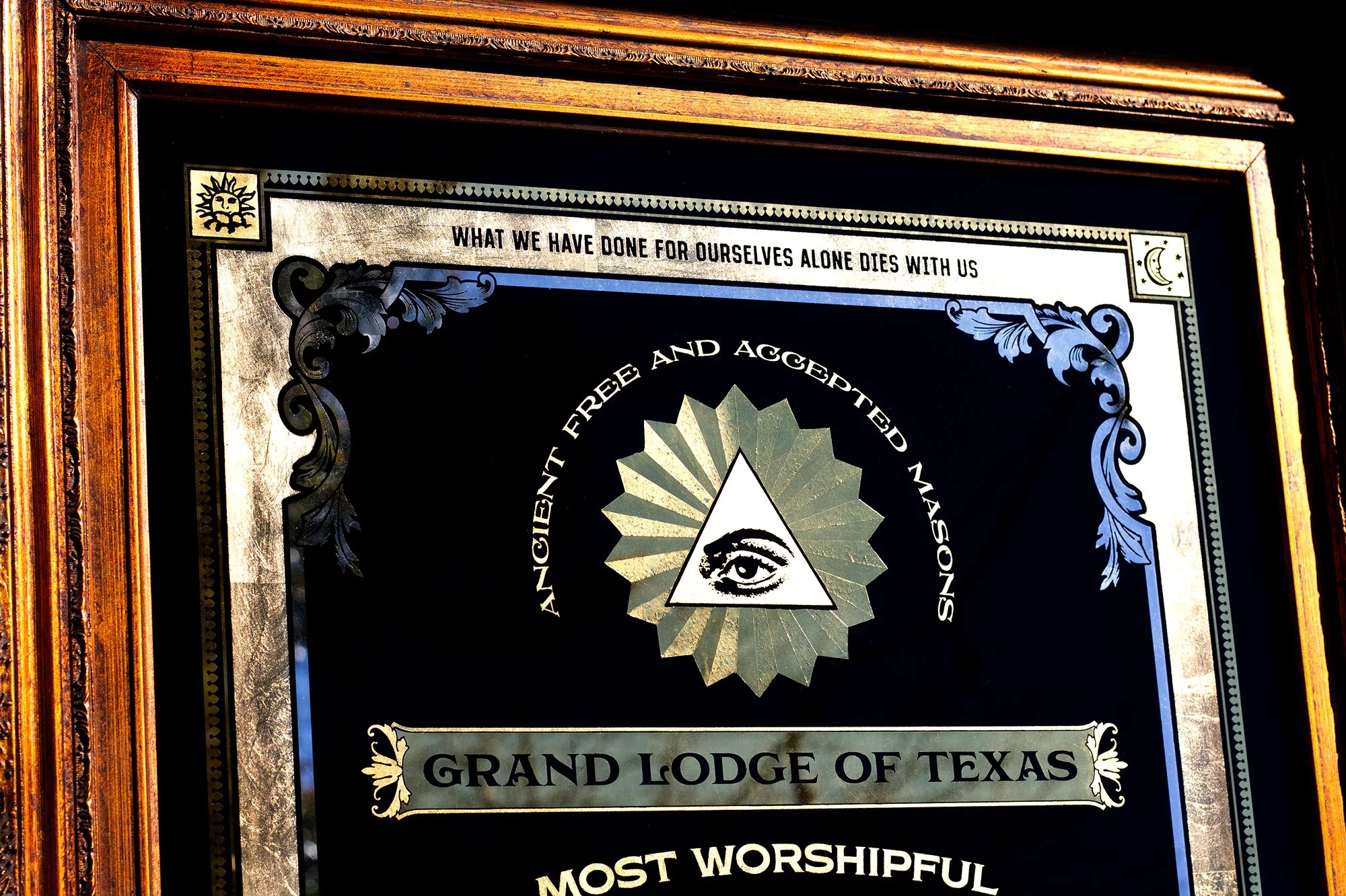 A framed decorative plaque with a black background and silver borders, featuring an eye within a triangle and the words "Ancient, Free, and Accepted Masons" around it. Text at the top says "What we have done for ourselves alone dies with us", and the