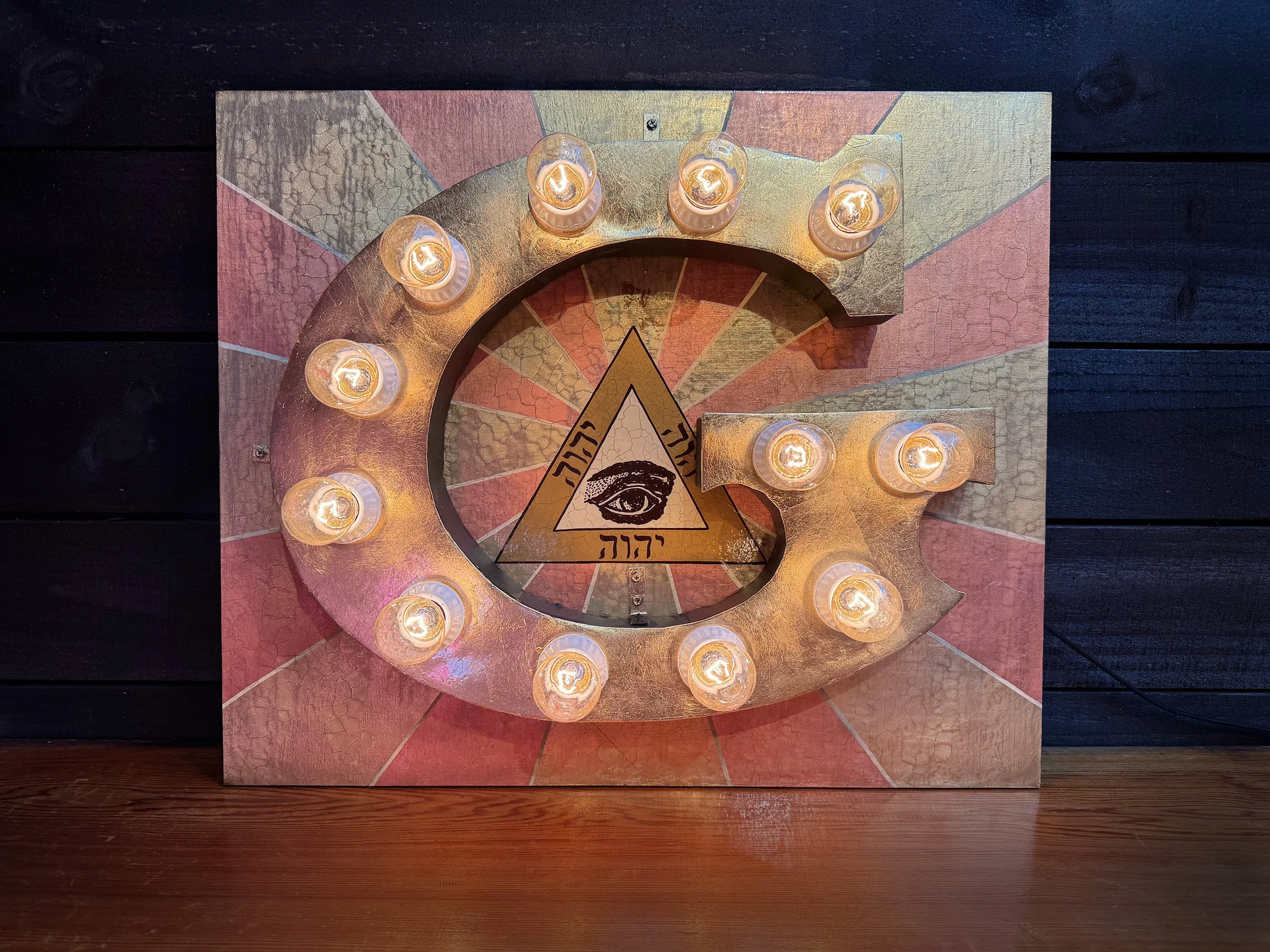 Decorative wall art featuring a golden letter G with string lights, a triangular symbol with an eye inside, and some Hebrew text.