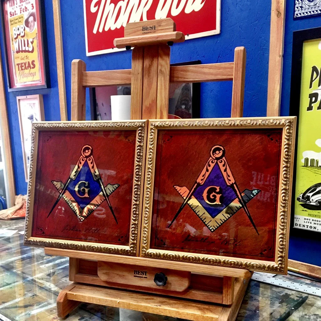 Two framed Masonic square and compass symbols with a letter G in the center, displayed on a wooden stand in front of a blue wall with vintage posters.