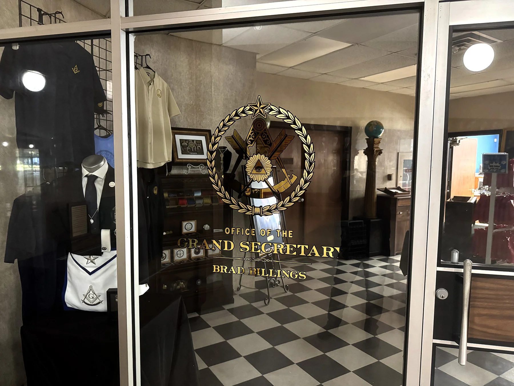 Glass door with a logo for the Office of the Grand Secretary, Brad Billings, featuring a square and compass symbol with a laurel wreath, inside an office with checkered black and white flooring, framed pictures, and displays of clothing and memorabil