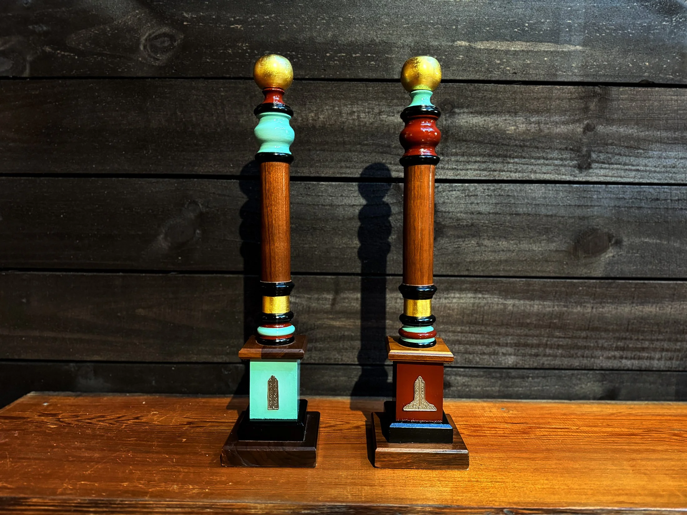 Two decorative wooden candlesticks with colorful accents and round top finials, placed on a wooden surface against a dark wood-paneled background.