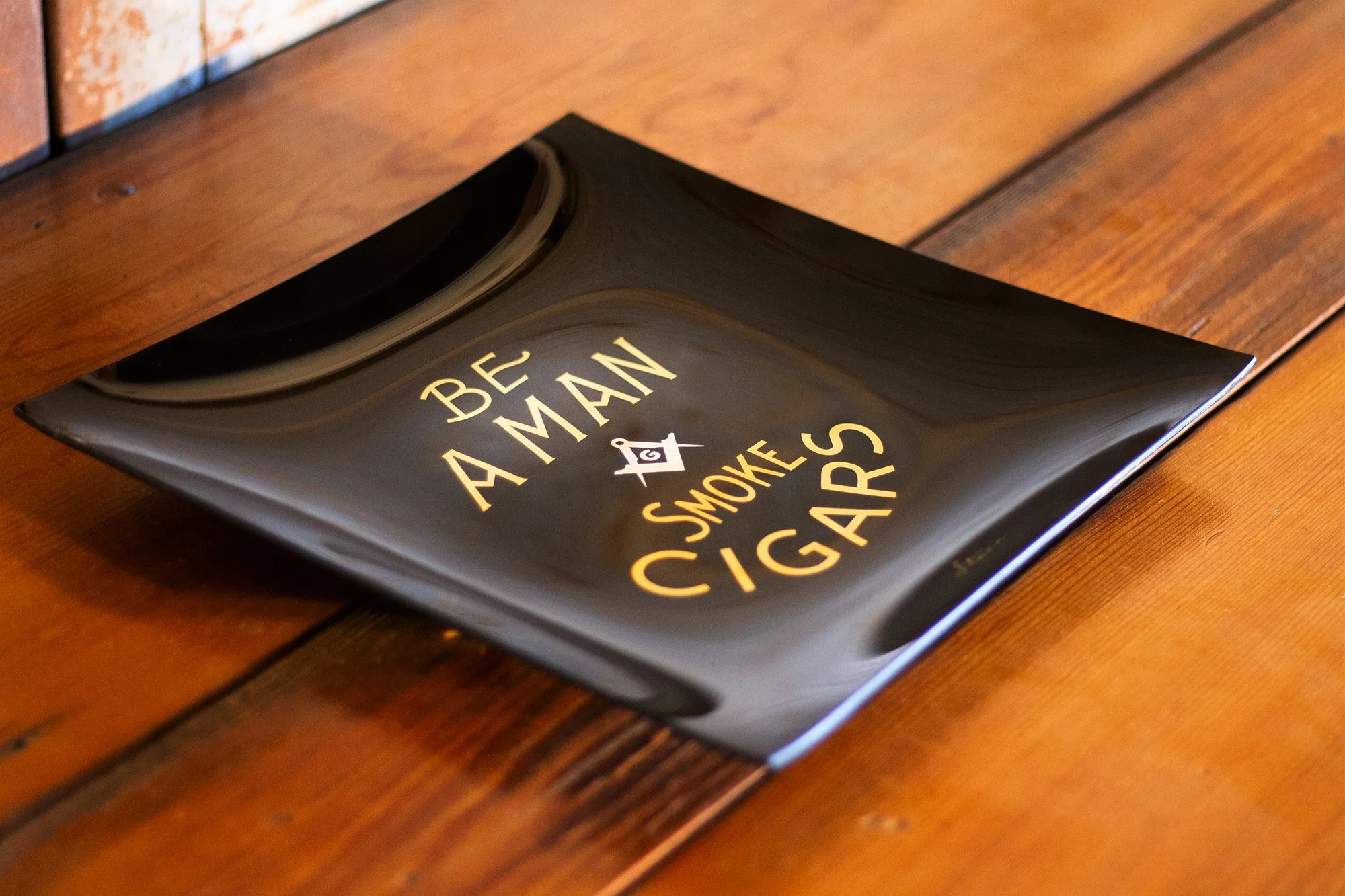 Square and Compasses Ashtray with the text 'Be A Man Who Smokes Cigars' in gold and white lettering, resting on a wooden surface.