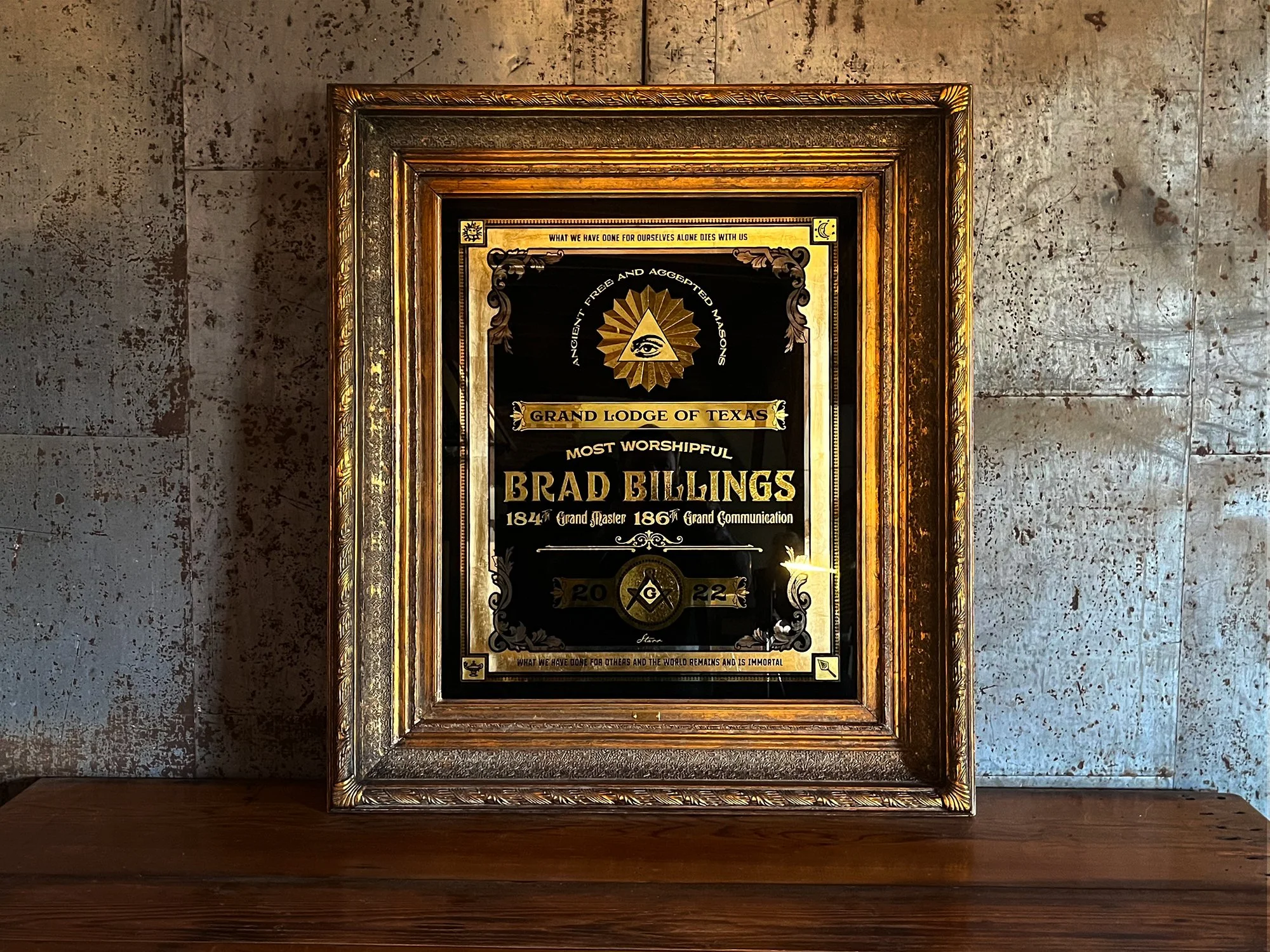 Framed certificate or plaque with gold and black design, reading 'Grand Lodge of Texas' and 'Brad Billings', with some symbols and dates, on a wooden table against a textured wall.