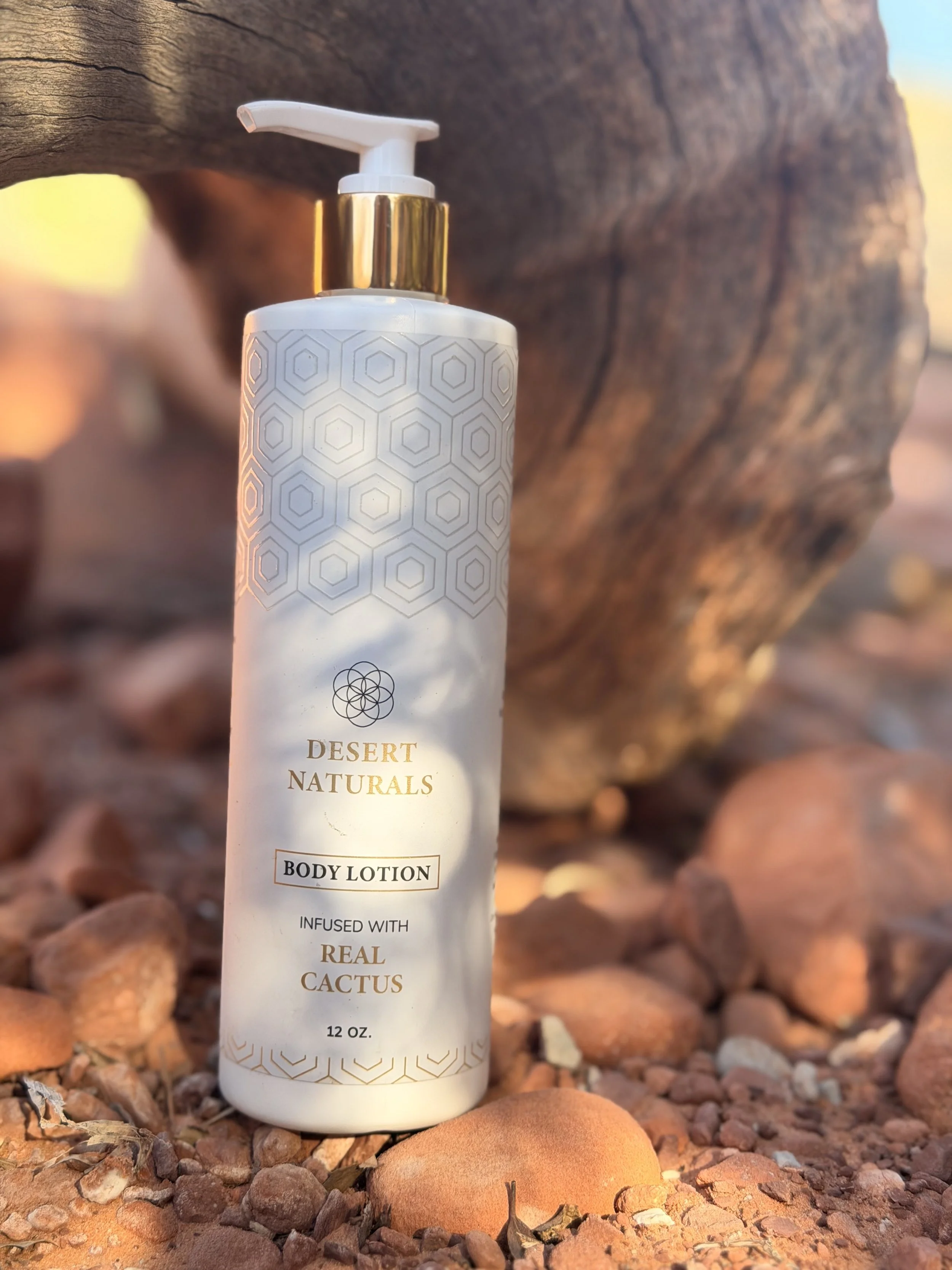 A white bottle of Desert Naturals body lotion with a pump dispenser, placed on rocky desert ground with a large rock in the background.
