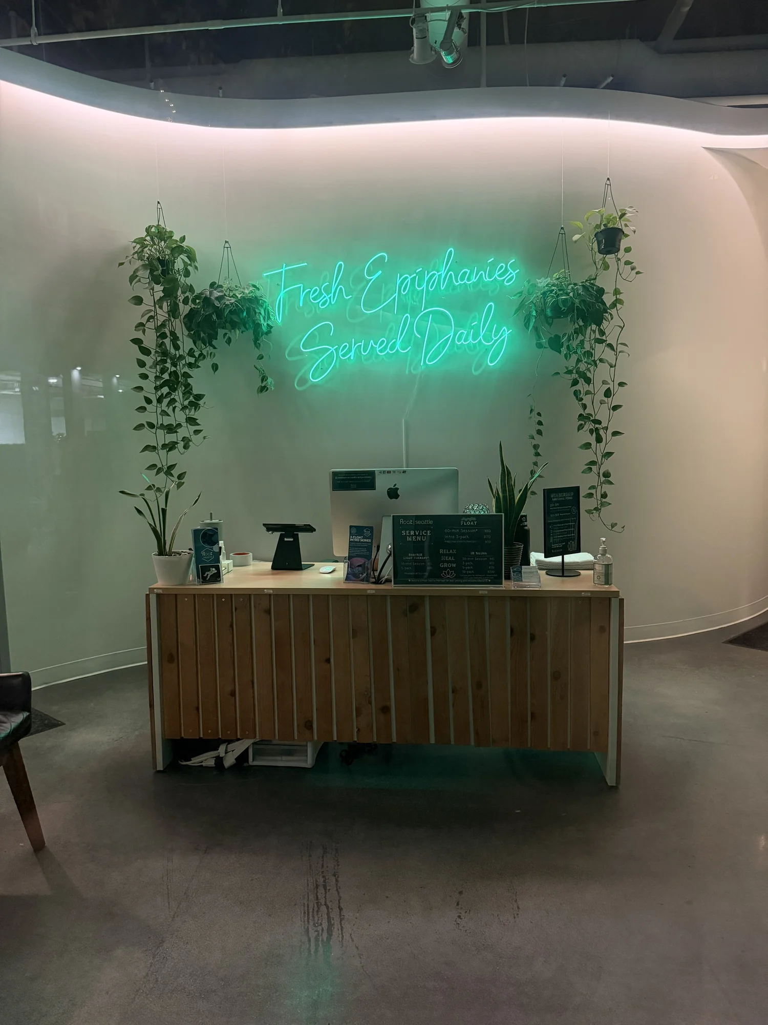 Front desk at the South Lake Union studio, with hanging plants and a green neon sign that reads Fresh Epiphanies Served Daily