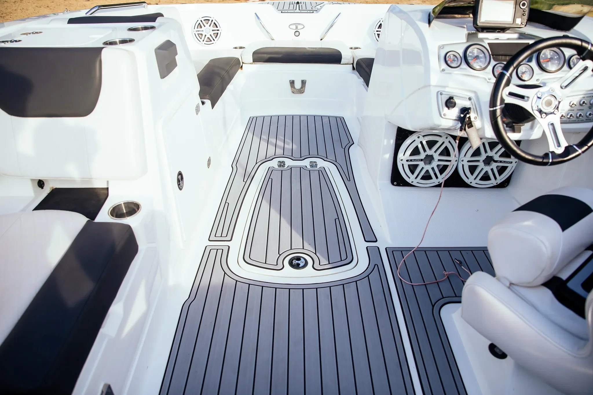 Empty boat interior with white and black seating, a black steering wheel, and multiple gauges and speakers on the dashboard, along with a gray and black non-slip floor mat.
