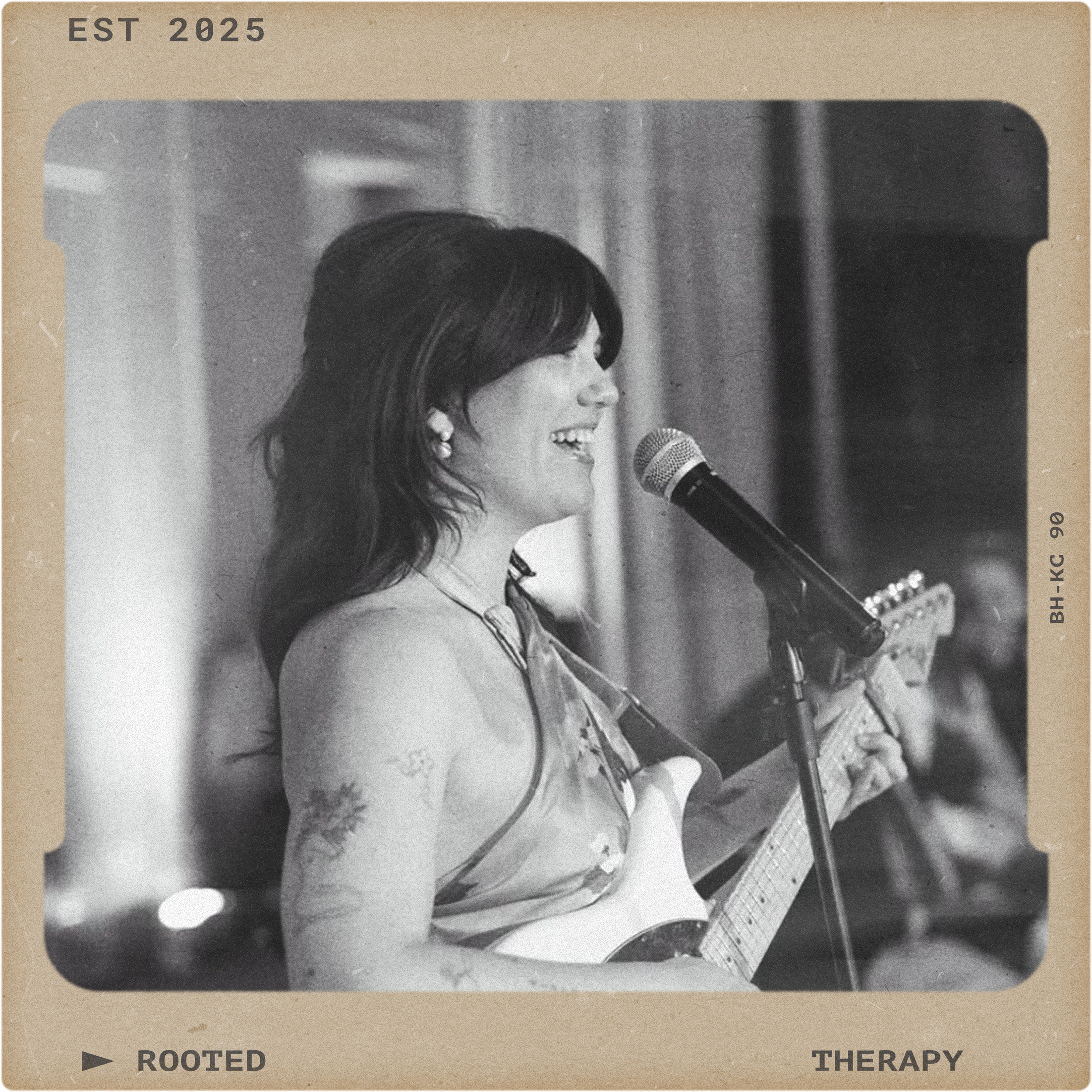 A woman with dark hair singing and playing an acoustic guitar into a microphone, smiling. She is wearing earrings and a sleeveless top. The background shows curtains, suggesting an indoor performance or therapy session, with a vintage or nostalgic tone.
