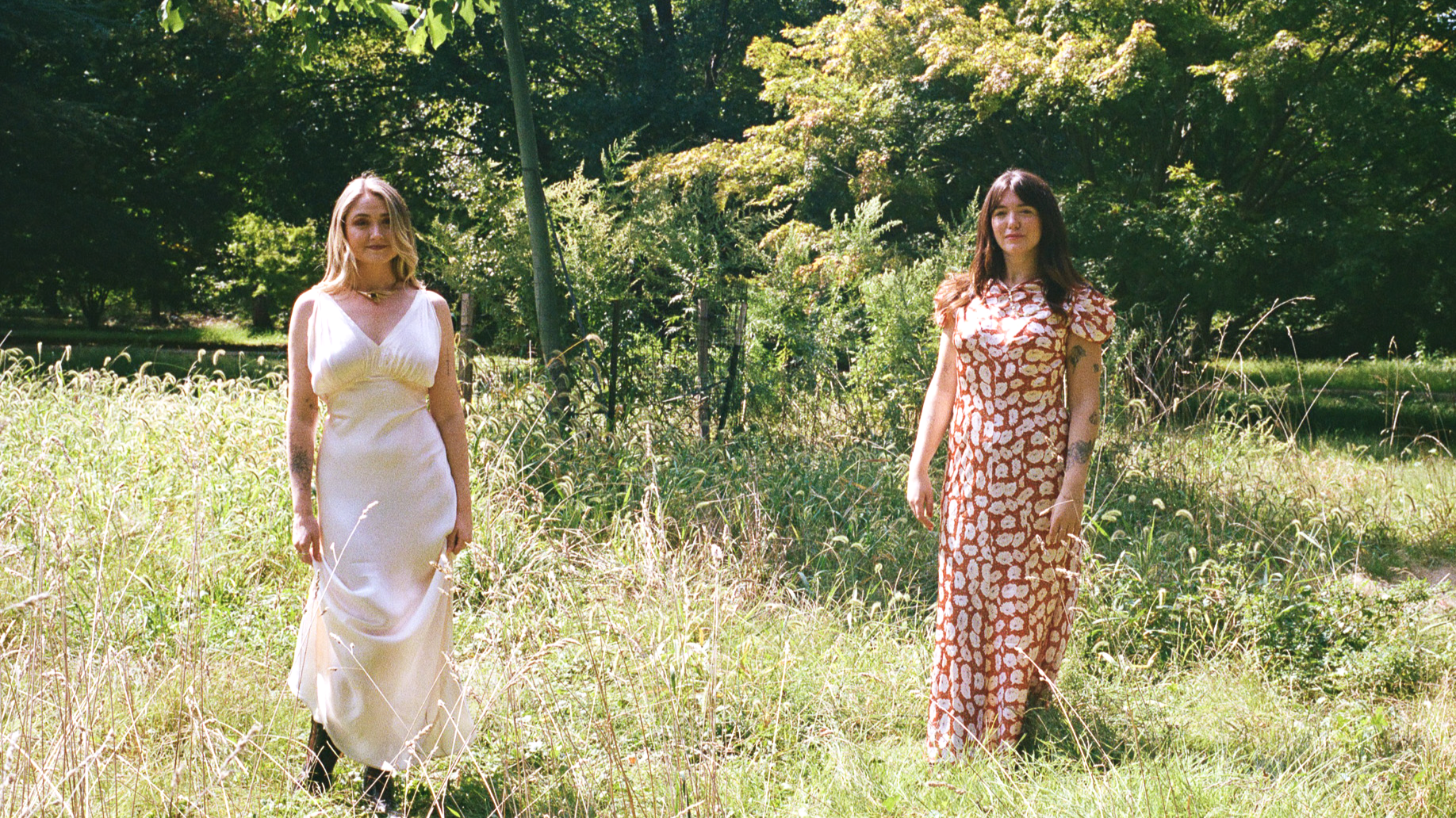 Two women standing in a grassy field with trees in the background, one wearing a white dress and the other in a patterned orange and white dress.