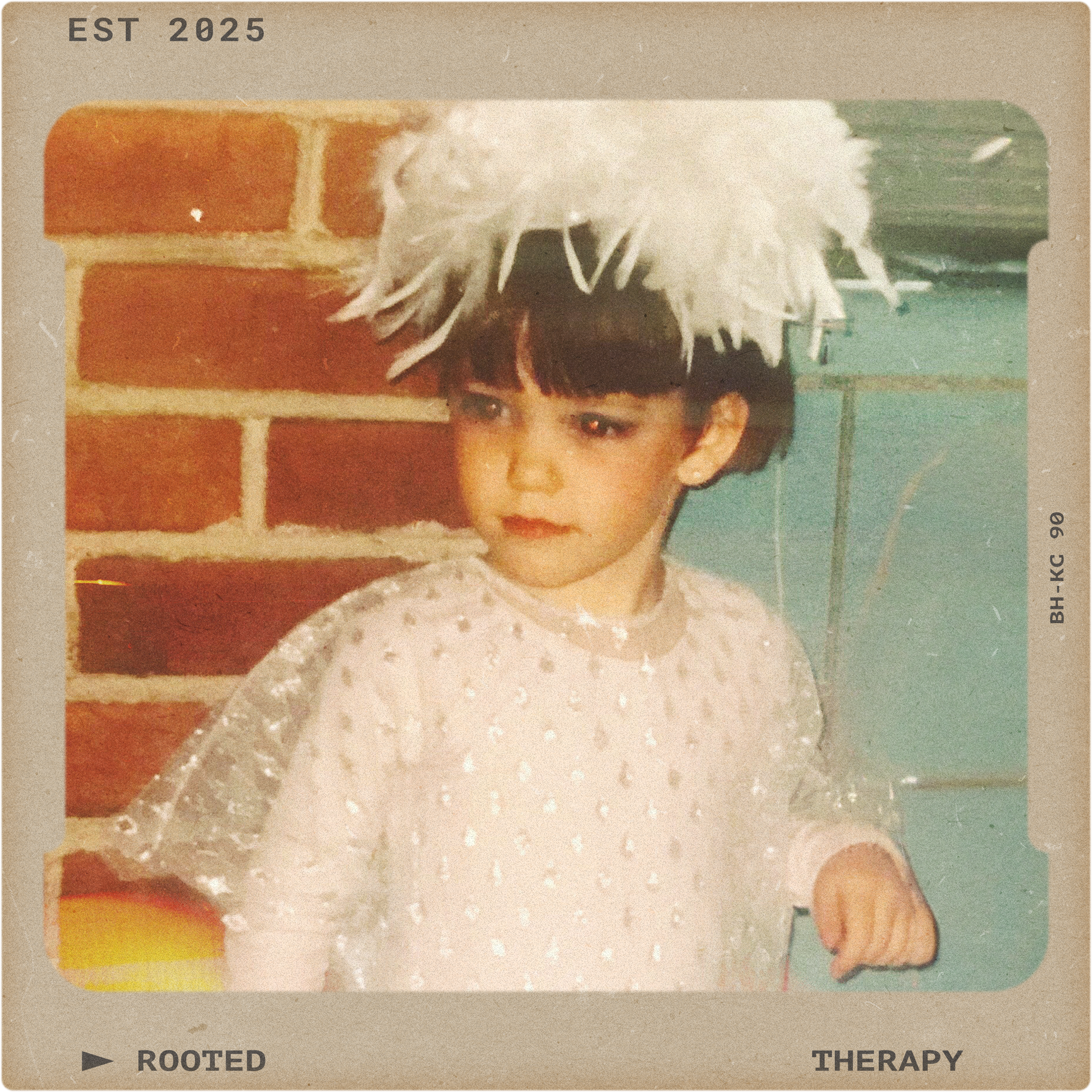 A young girl wearing a white dress with shiny details and a fluffy white headpiece, sitting against a brick wall background.
