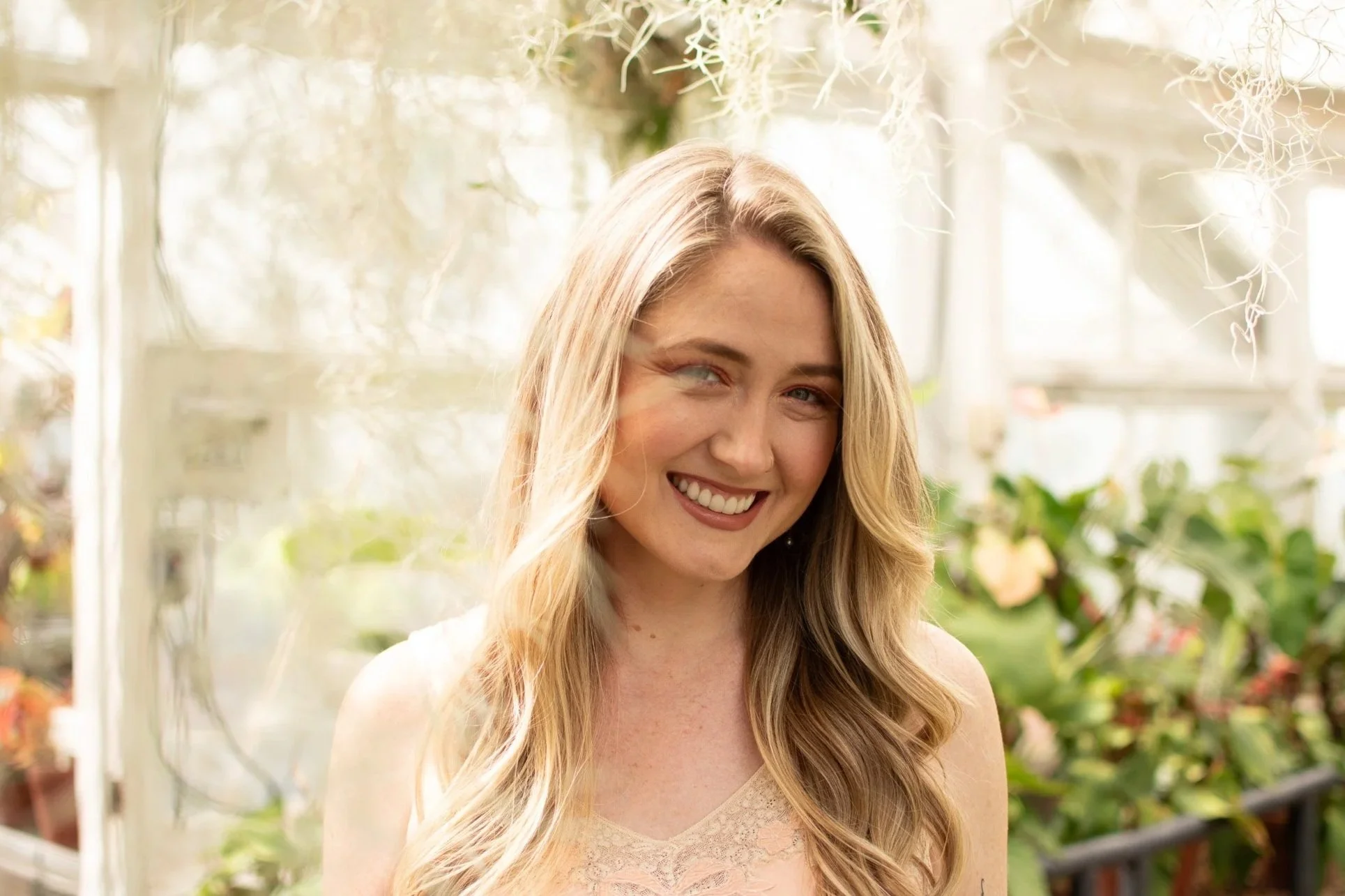A smiling woman with long blonde hair in a greenhouse with plants in the background.