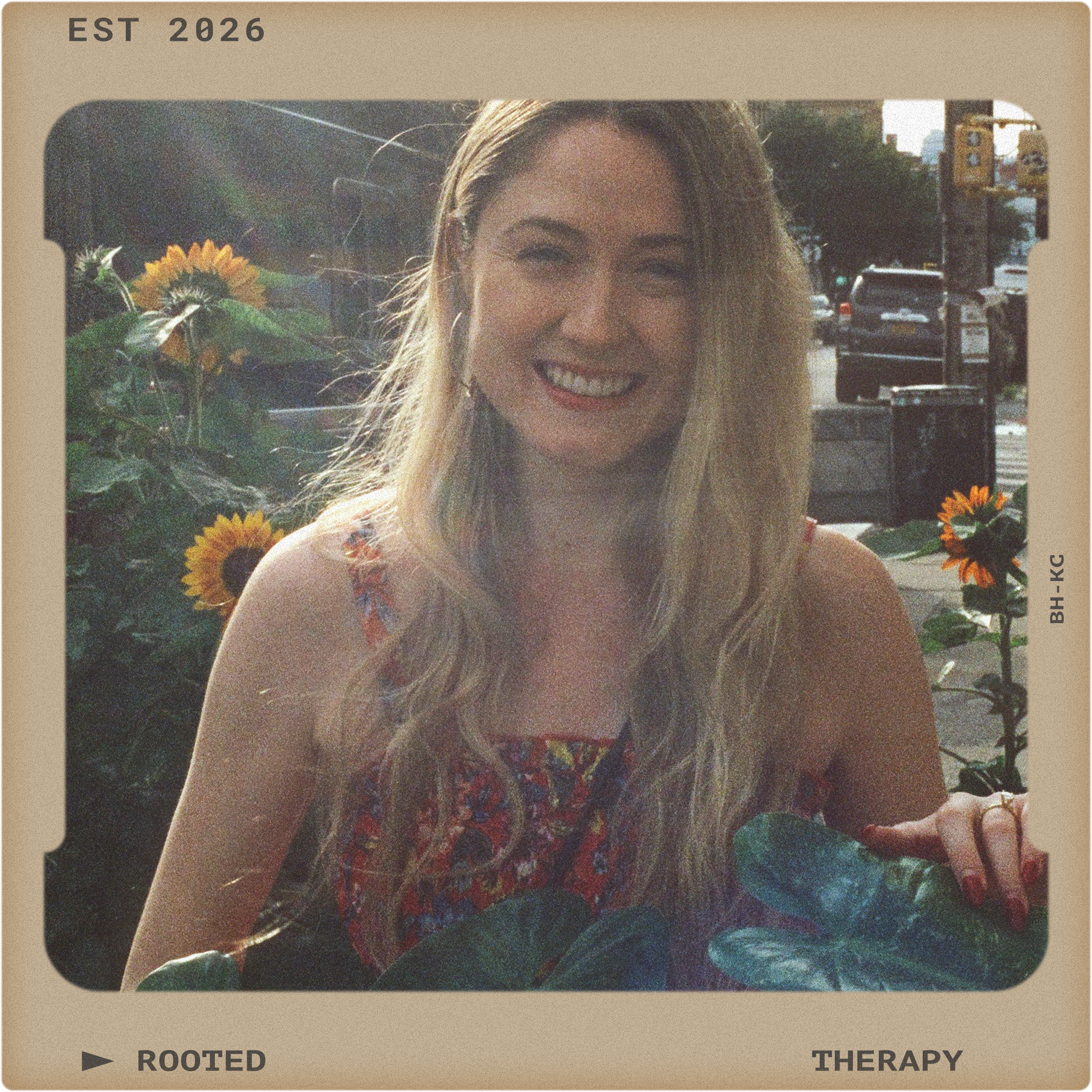 A woman with long blonde hair smiling, sitting outdoors with sunflowers and green plants, in an urban setting.