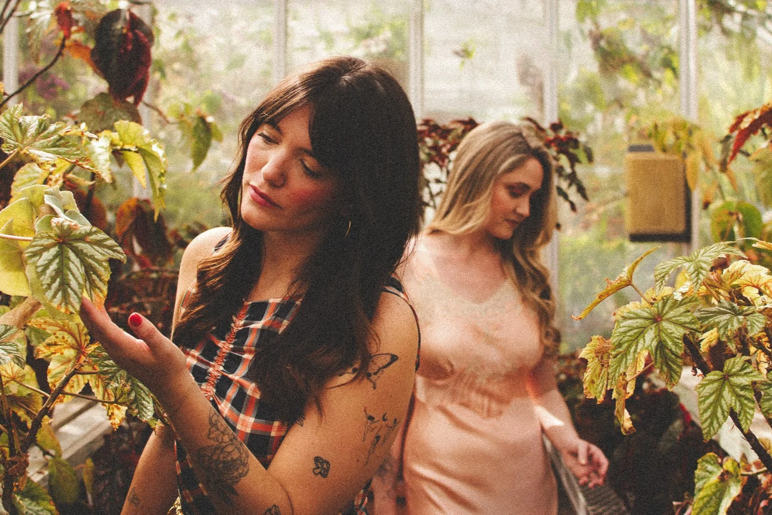 Two women with tattoos and long hair in a greenhouse surrounded by green and red plants, one is touching a leaf and the other is looking down.
