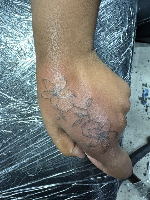 A tattoo outline of a flower with leaves on a person's hand.
