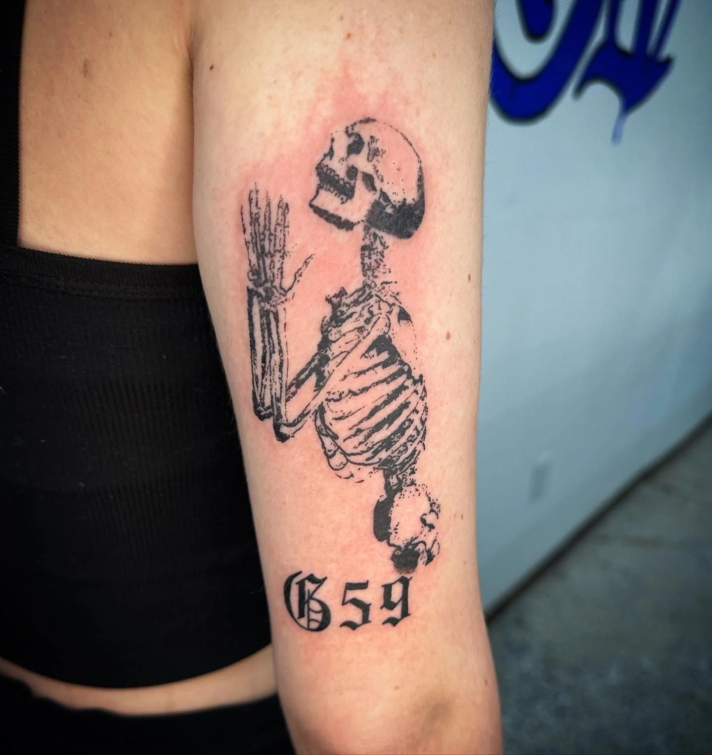 Tattoo of a skeleton with a hand raised, number 'g59' in black gothic font, on person's upper arm.