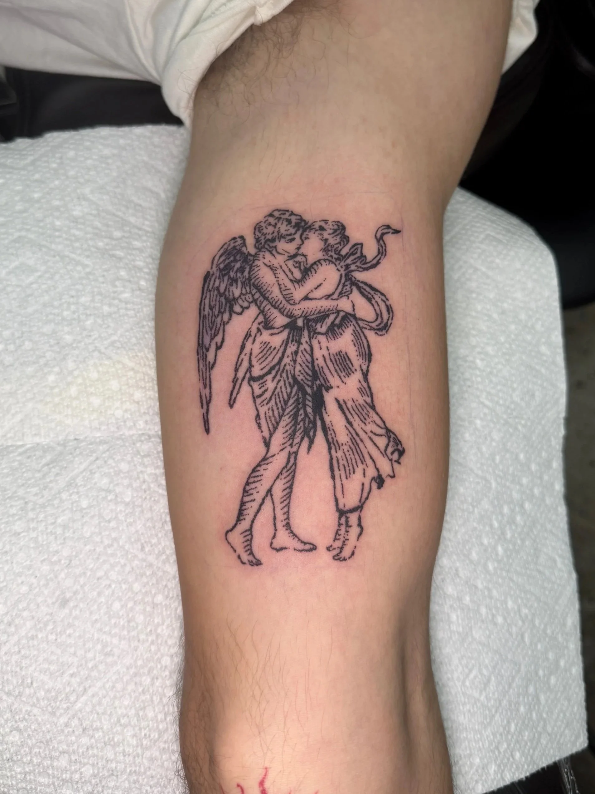Tattoo of a classical-style drawing of two angels embracing, with one angel having wings, on a person's calf.