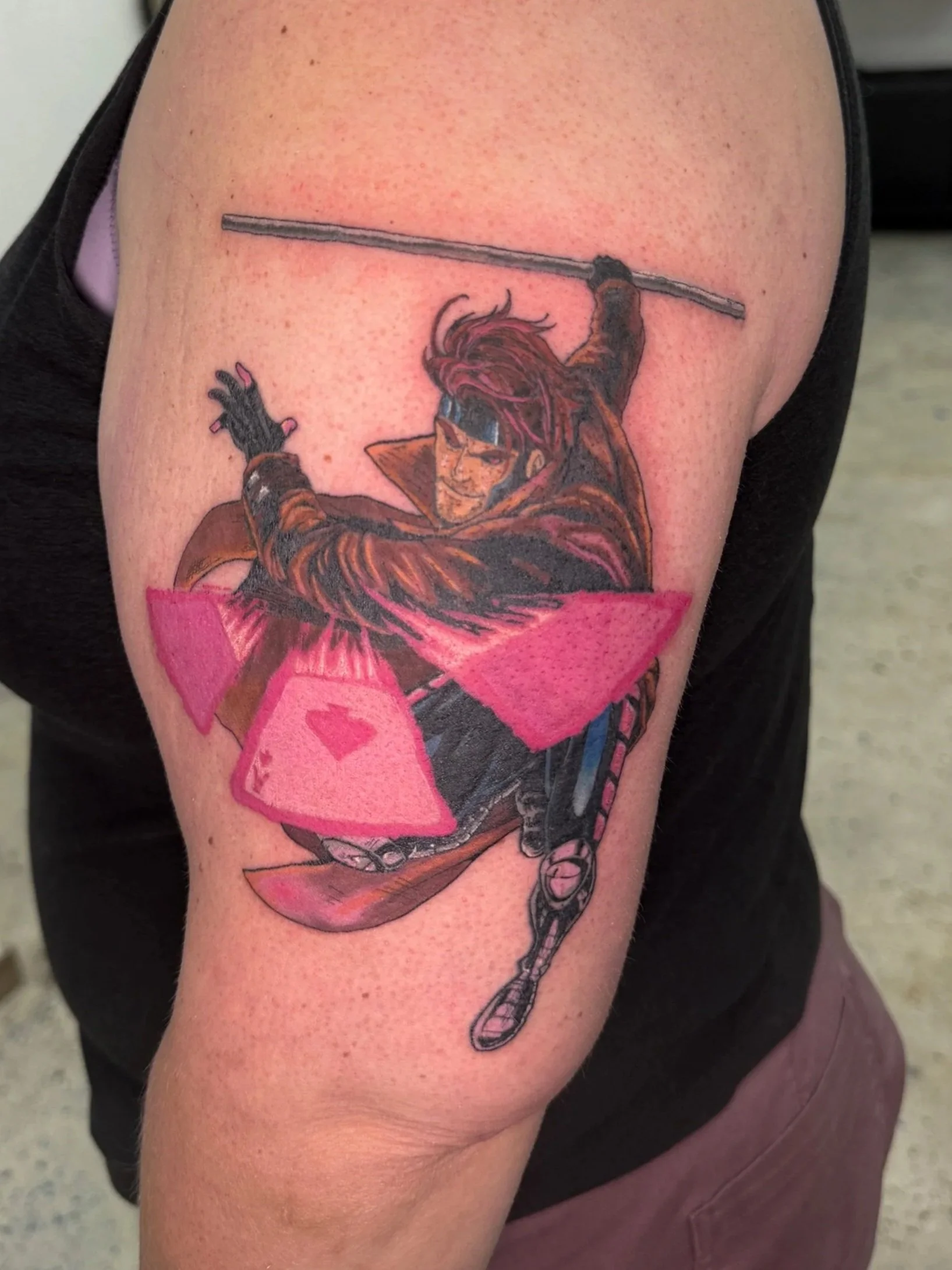 Tattoo of a male character in a dynamic pose with a staff, wearing sunglasses and a brown coat, slicing through pink geometric shapes on the upper arm.