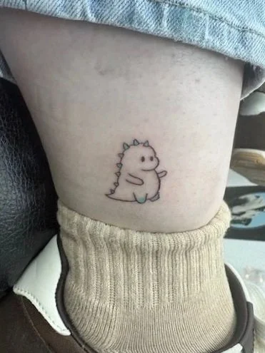 Tattoo of a cute, smiling cartoon dinosaur with spikes on its back on a person's upper arm.