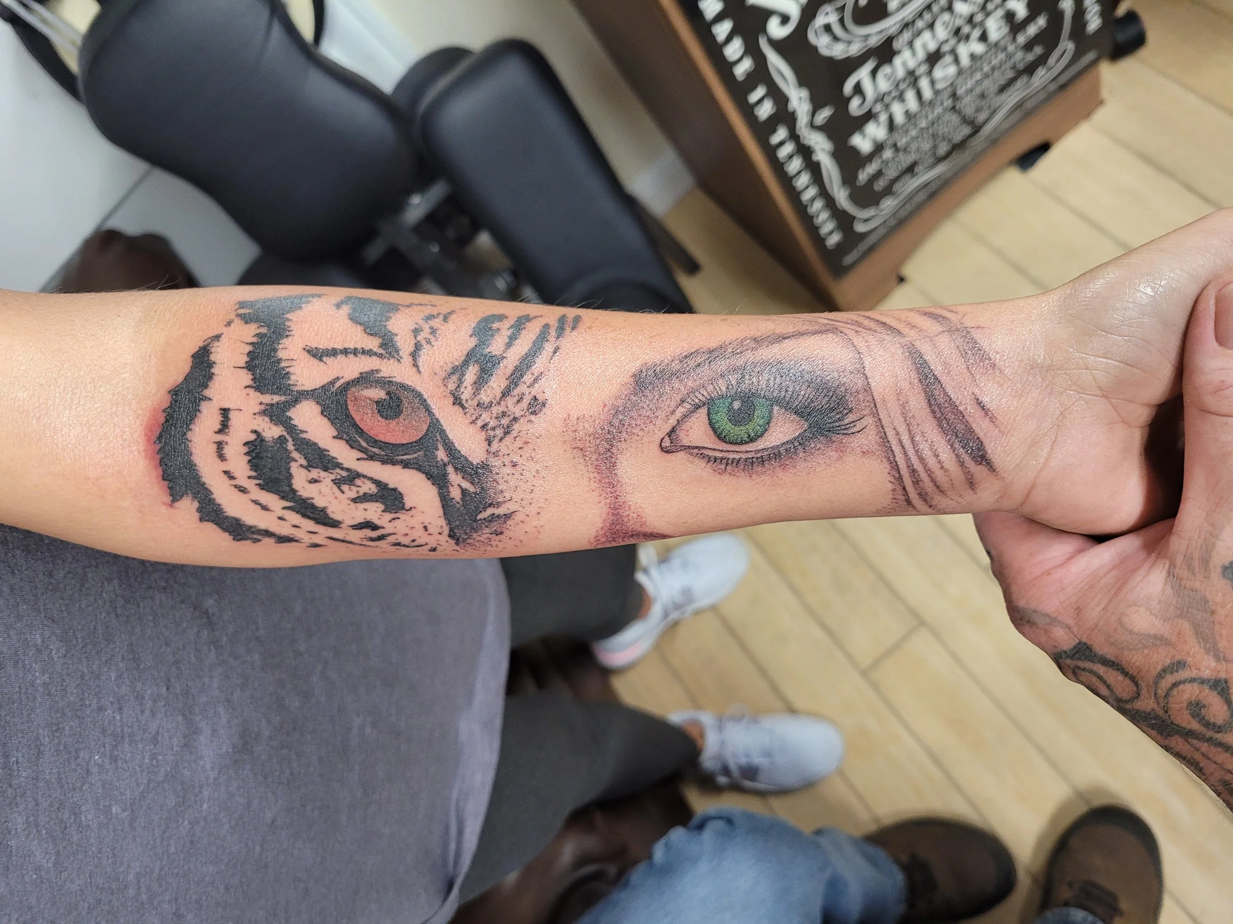 A tattoo on a person's forearm depicting two eyes, one detailed realistically with a green iris and the other abstract with black lines and a brown iris.