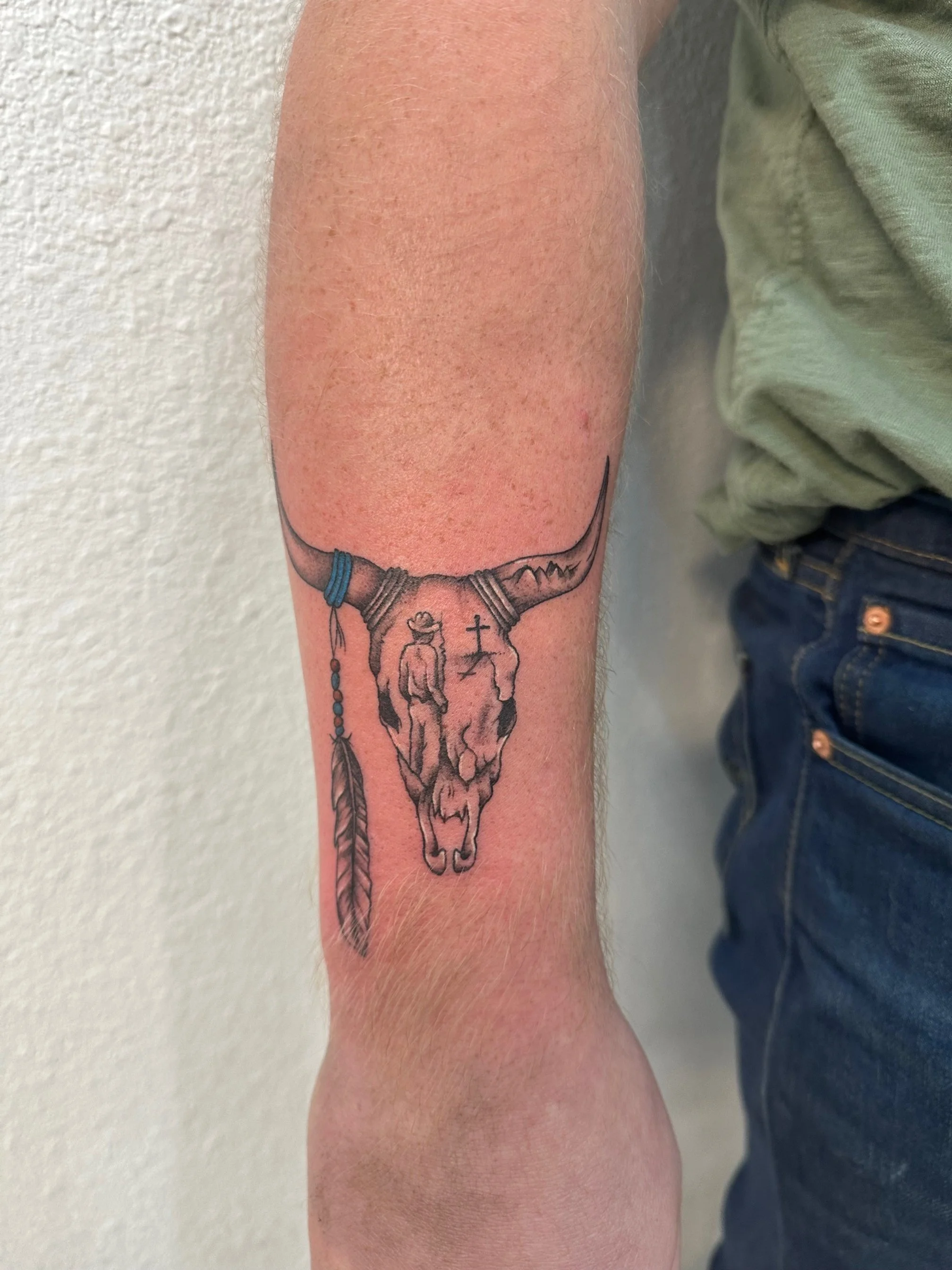 Tattoo of a bull skull with feathers and beads, and a small cross between the horns, on a person's forearm.