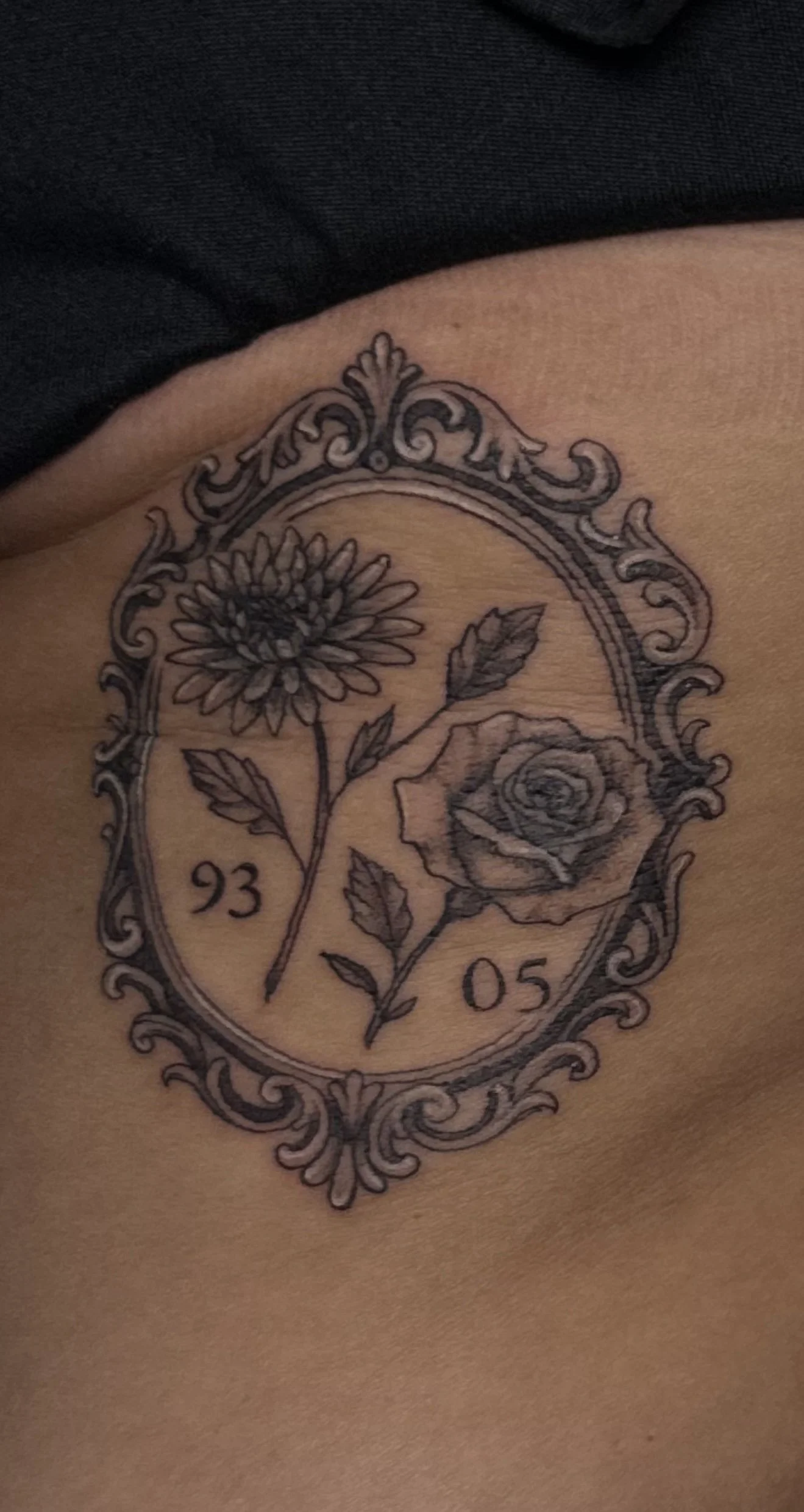 Tattoo of a floral design with a daisy and a rose inside an ornate oval frame, with the numbers 93 and 05.