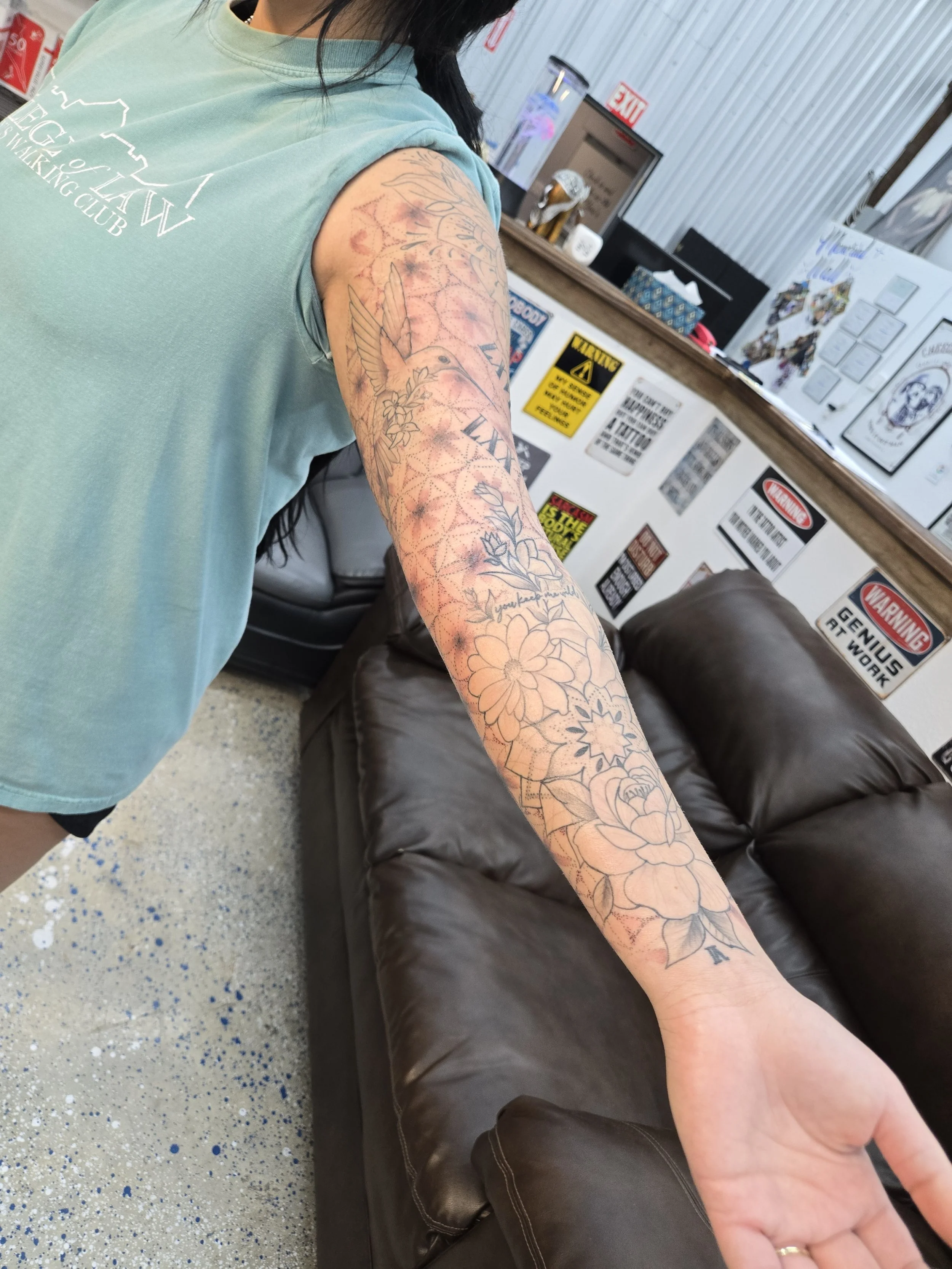A person with a long floral tattoo sleeve on their arm, wearing a light green t-shirt, standing in a waiting area with a brown leather couch and a wall decorated with various signs and posters.