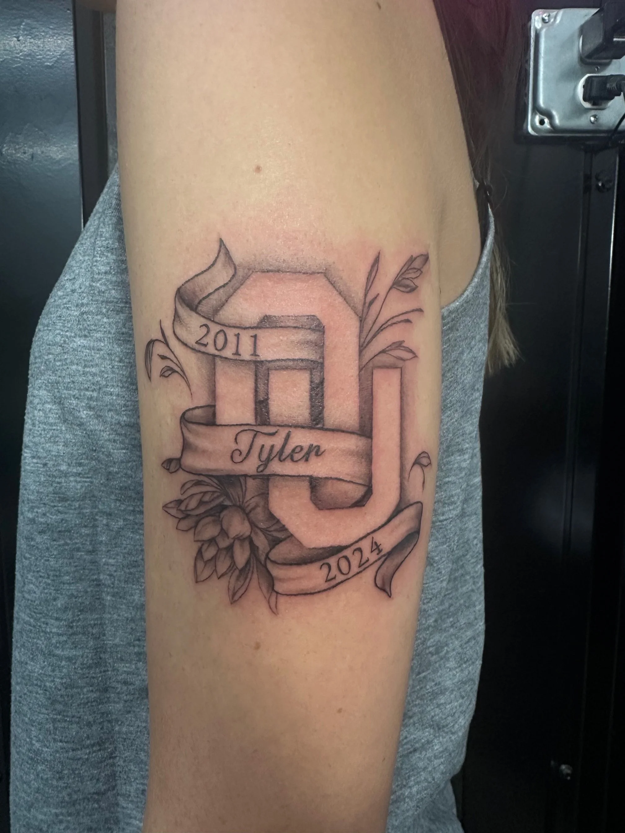 Tattoo of the letter J with a ribbon banner that says 'Tyler', surrounded by leaves, with the years 2011 and 2024, on a person's upper arm.