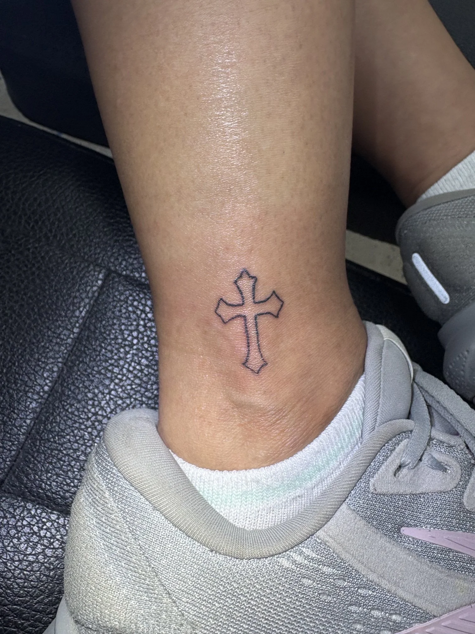 A tattoo of a cross with decorative patterns on a person's ankle.