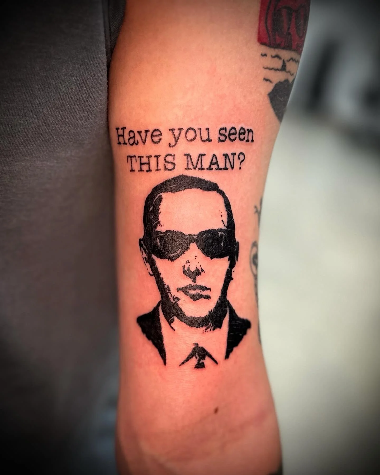 Tattoo of a man's face wearing sunglasses and a collared shirt, with the text 'Have you seen THIS MAN?'