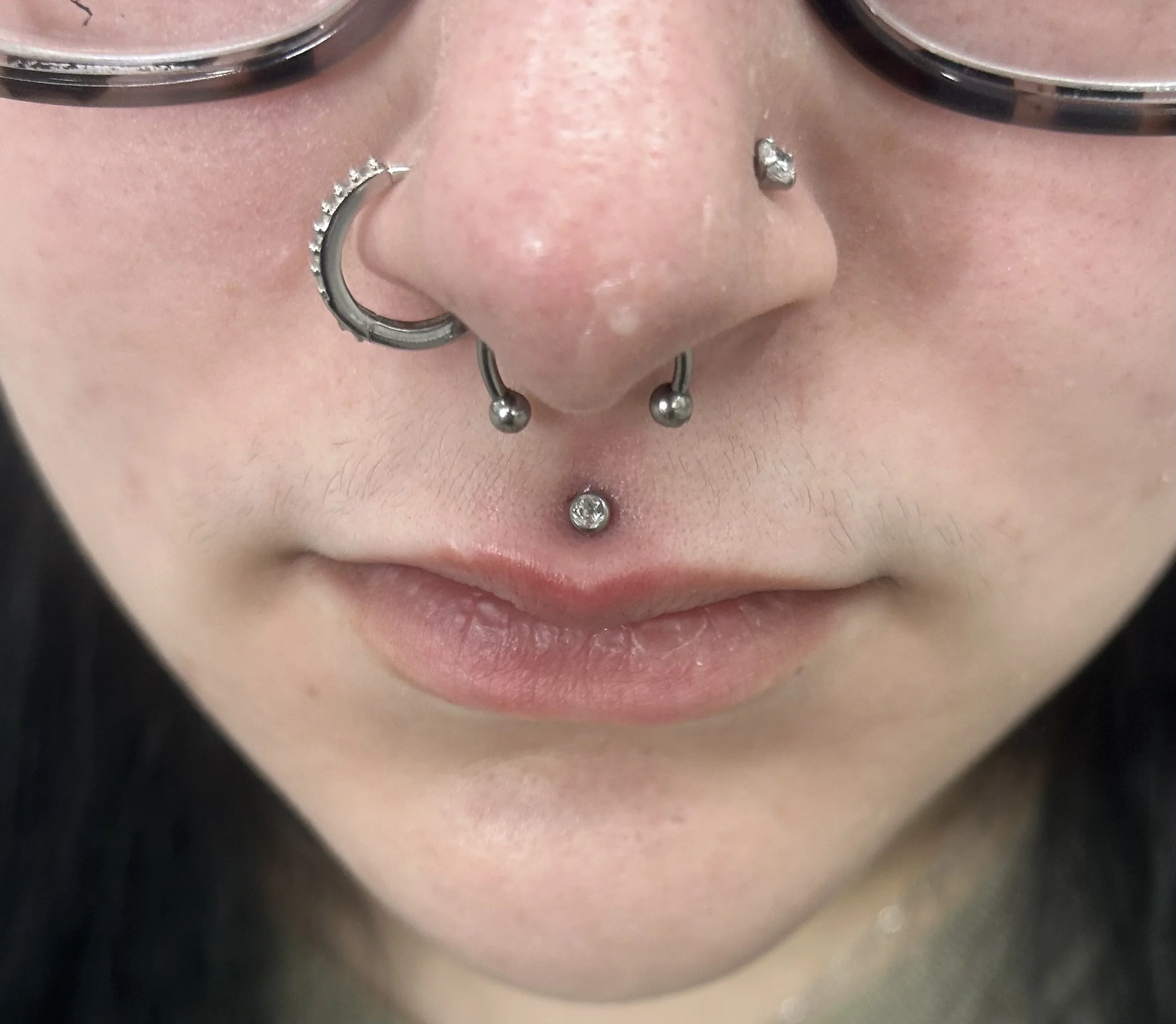 Close-up of a person's nose and mouth showing multiple facial piercings, including a septum piercing with a hoop, a nostril piercing, two upper lip piercings with small gemstones, and a labret piercing with a gemstone.