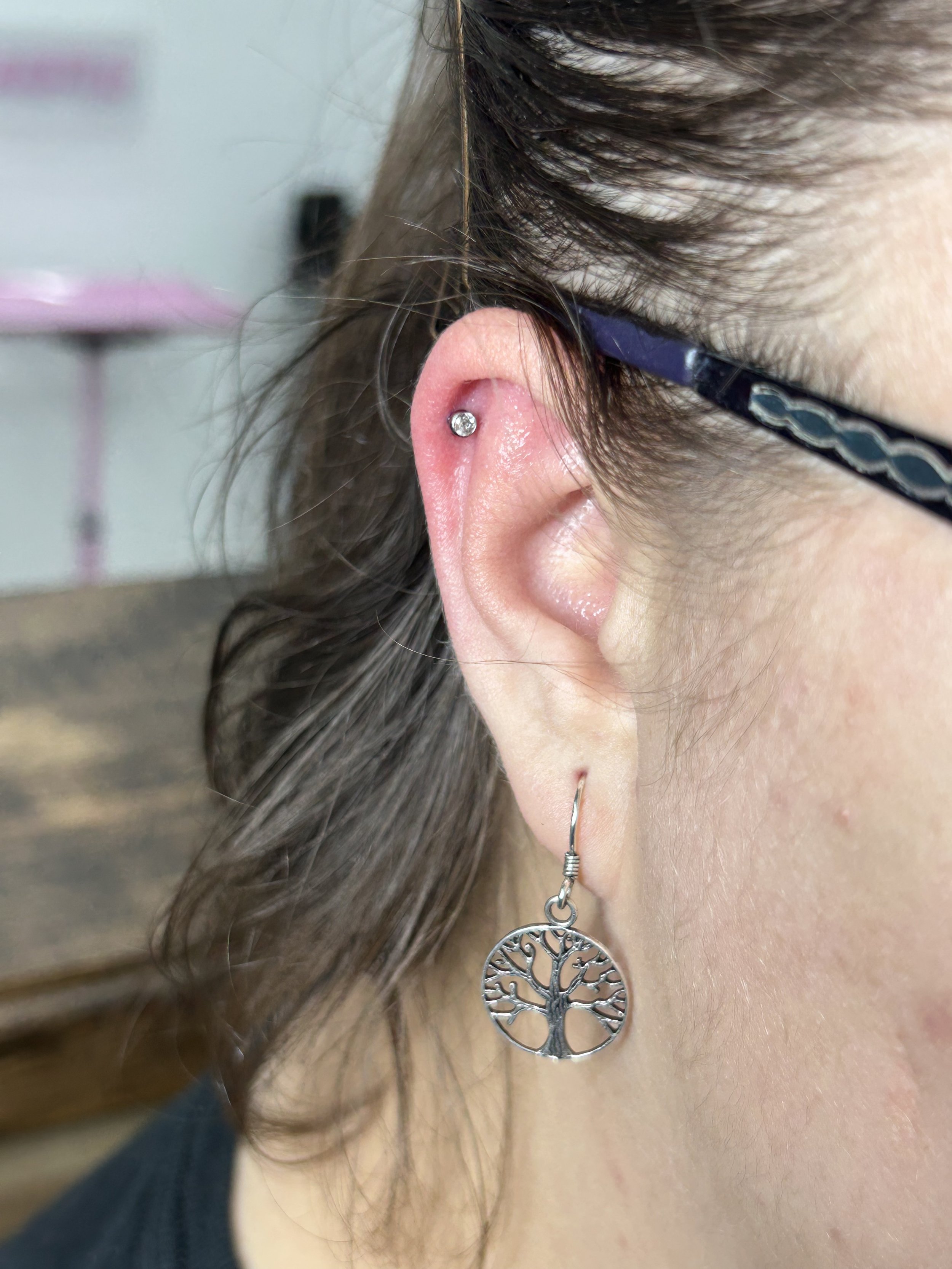 Close-up of a woman's ear with a small hoop earring and a dangling silver tree of life pendant earring, wearing glasses, with brown hair.