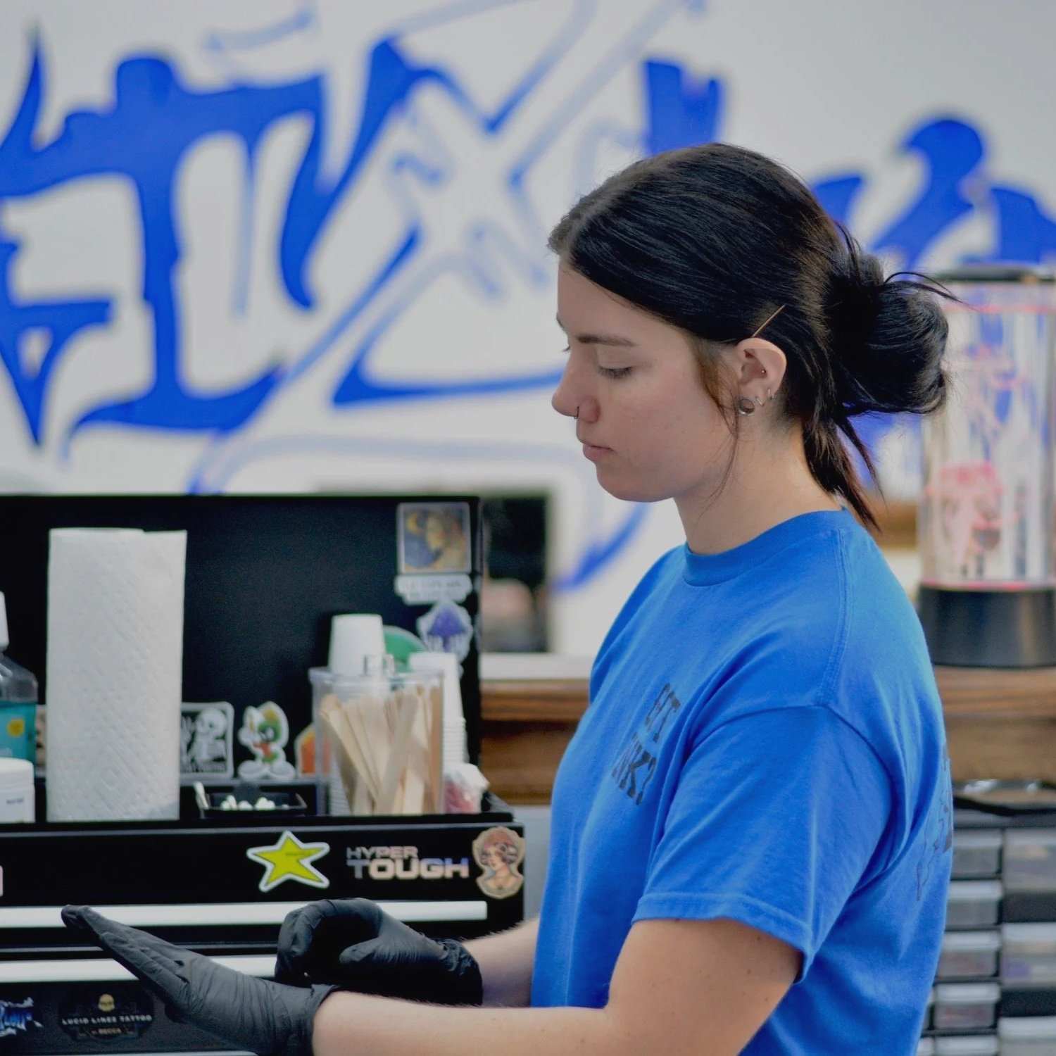 A young woman with dark hair tied back, wearing a blue T-shirt and black gloves, standing at a countertop in a store or tattoo parlor, looking down at a tablet or phone. Behind her are various objects and a wall with large stylized blue graffiti.