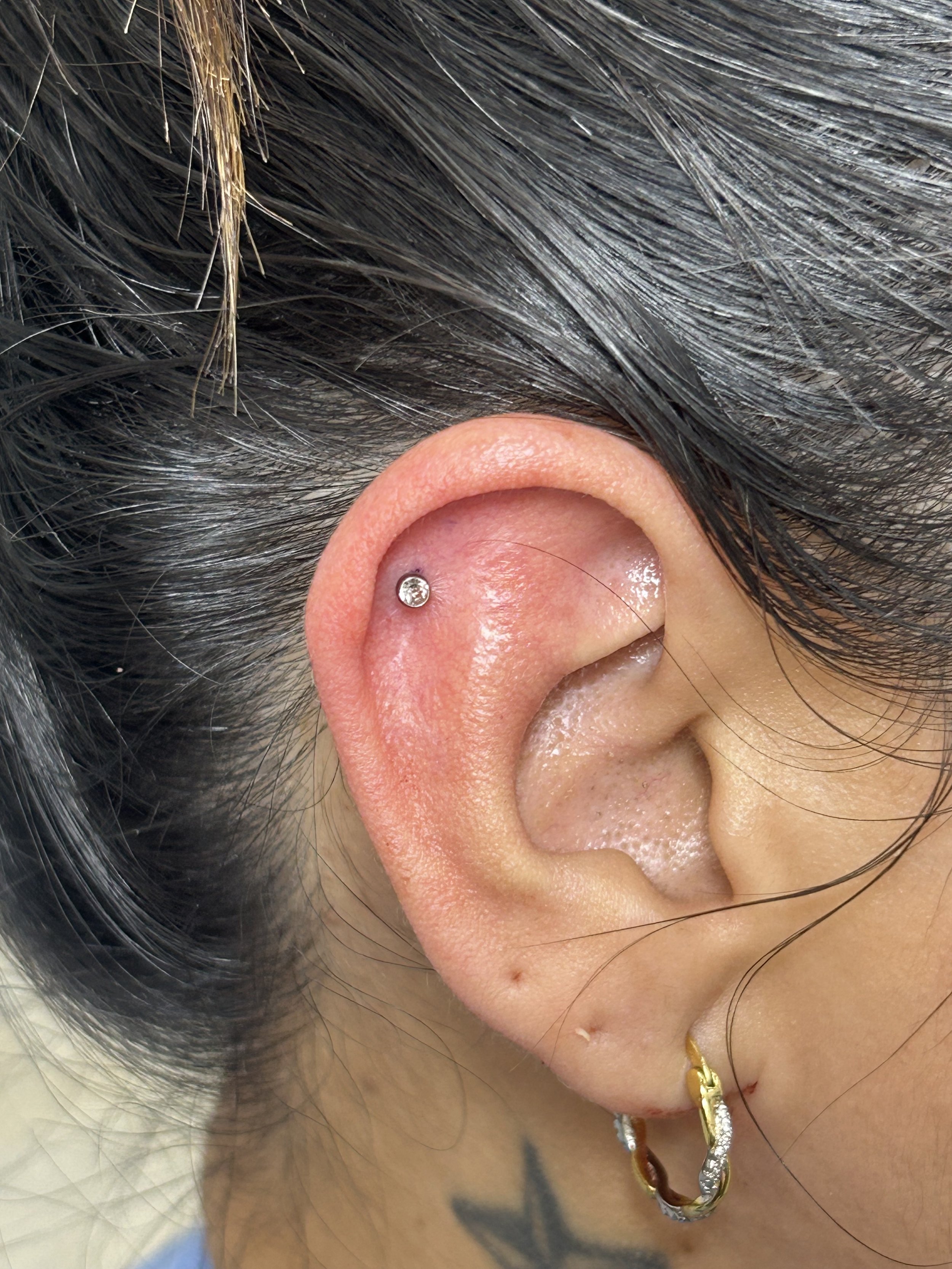 Close-up of a person's ear with a small silver hoop earring, dark hair, and a gold chain necklace visible.
