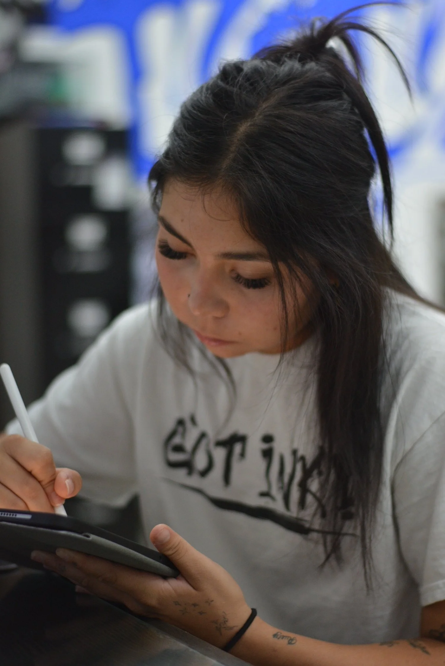 A young woman with long dark hair in a ponytail appears to be drawing or writing on a tablet or digital device at a desk, with a blurred background of supplies or objects.